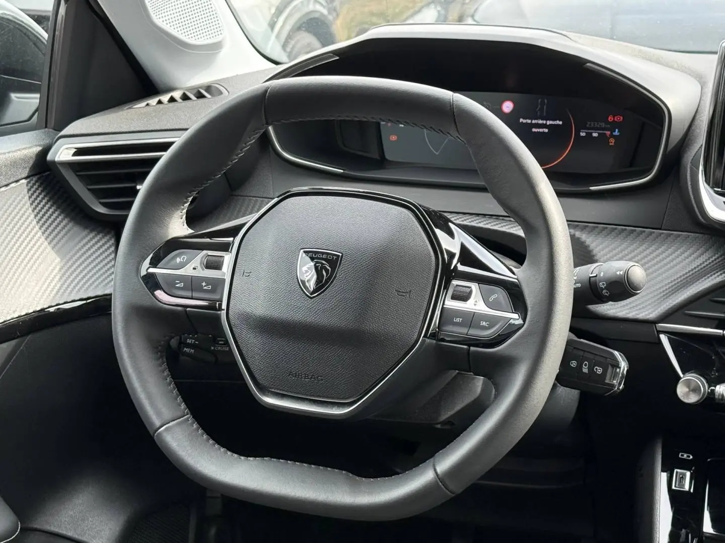 Vue rapprochée du volant noir de la Peugeot 208 2025 avec tableau de bord digital et détails carbone autour.