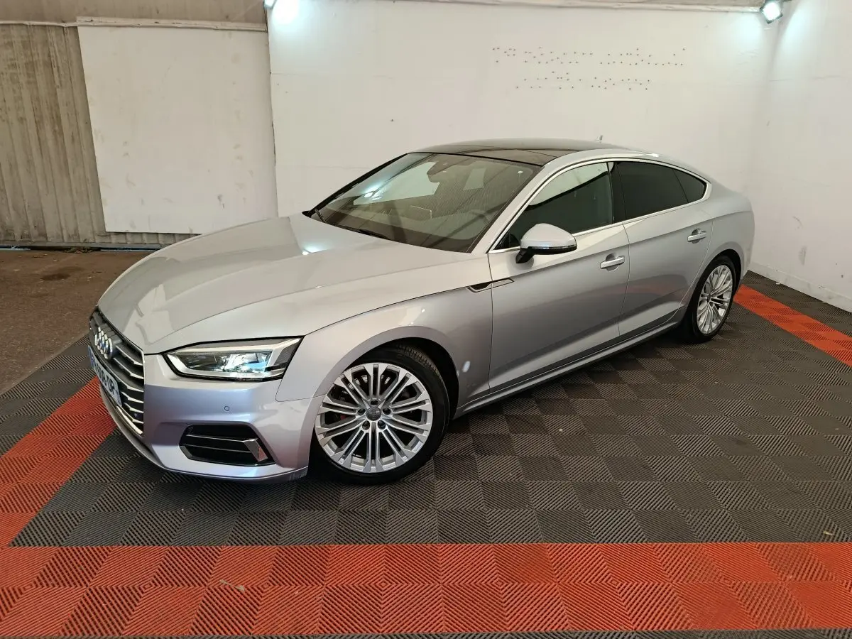 Audi A5 gris clair vue 3/4 avant droit dans un garage avec sol noir et rouge, jantes alliage multibranches.