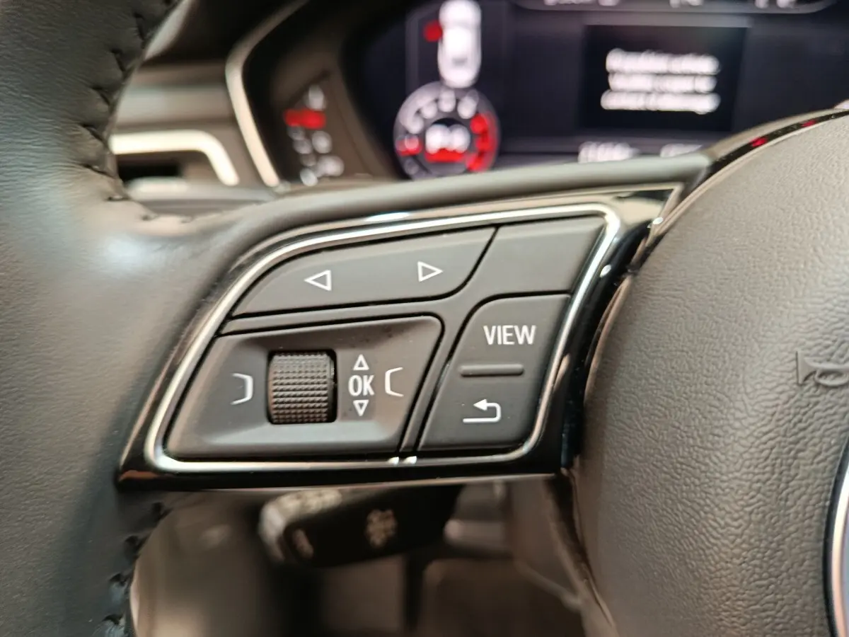 Gros plan sur les commandes multifonctions côté gauche du volant d’un Audi A5 gris clair, tableau de bord flou en arrière-plan.
