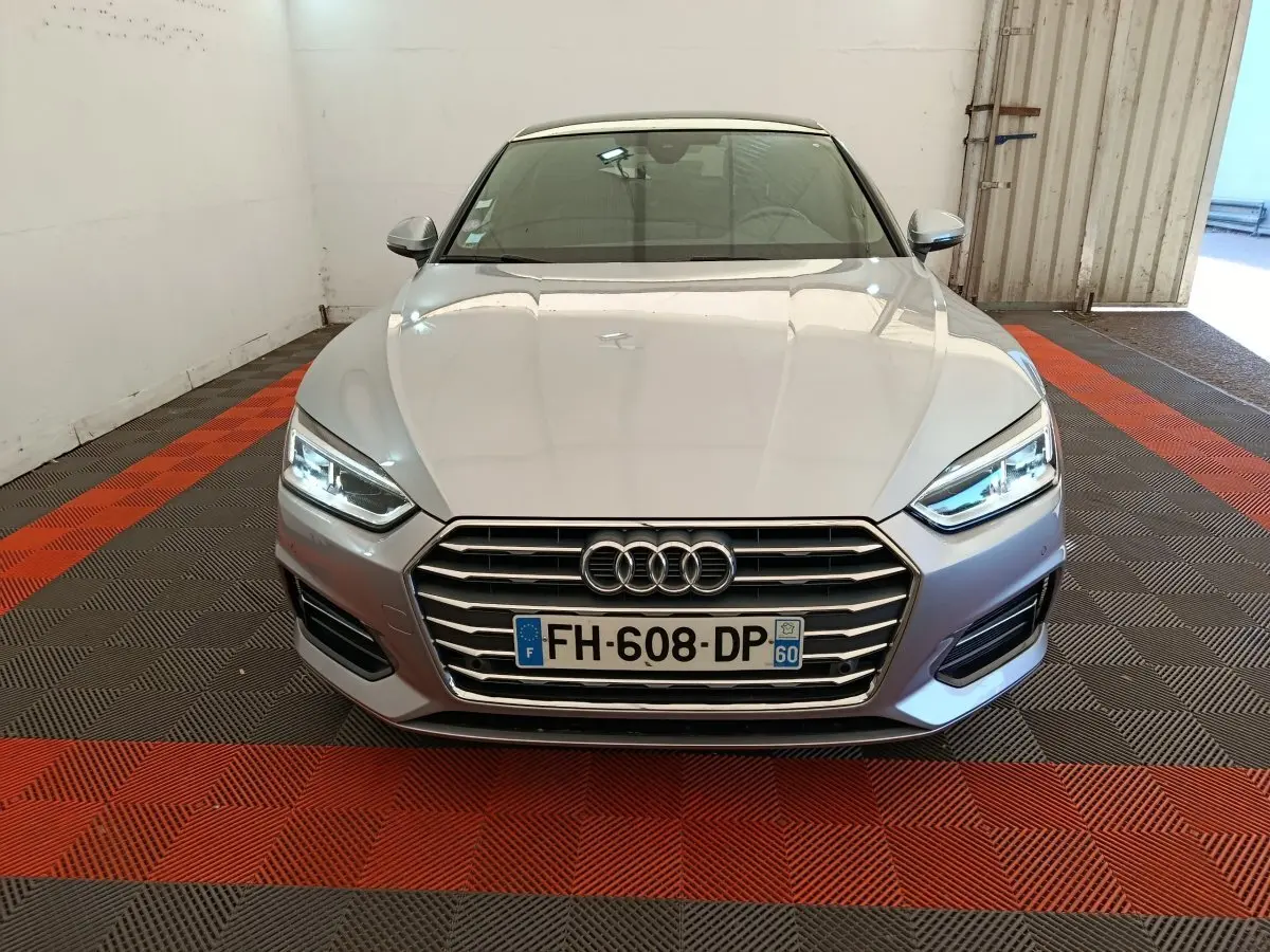 Vue frontale d'une Audi A5 gris clair avec phares LED allumés et calandre chromée dans un garage.