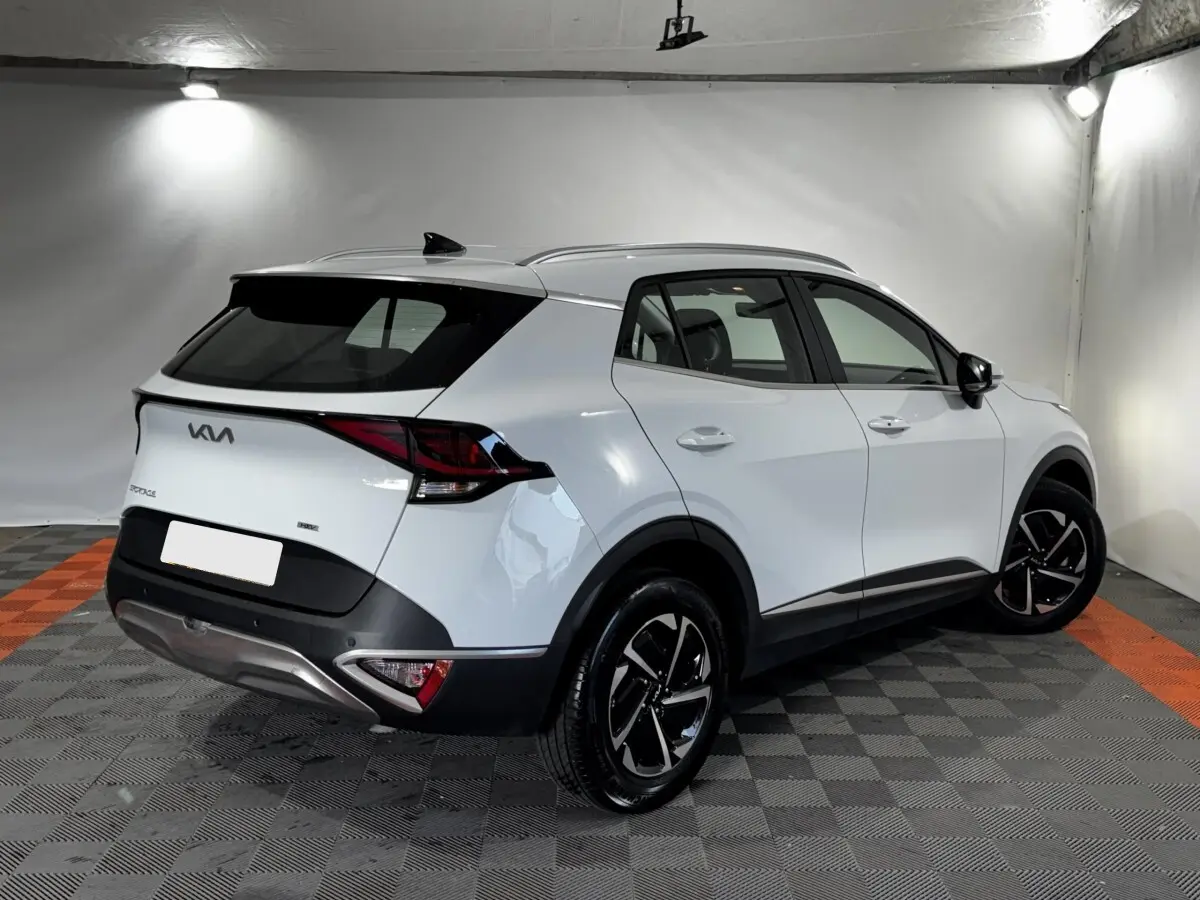 Vue 3/4 arrière droite d'un Kia Sportage blanc 2024 avec jantes noires et toit noir contrasté dans un studio.