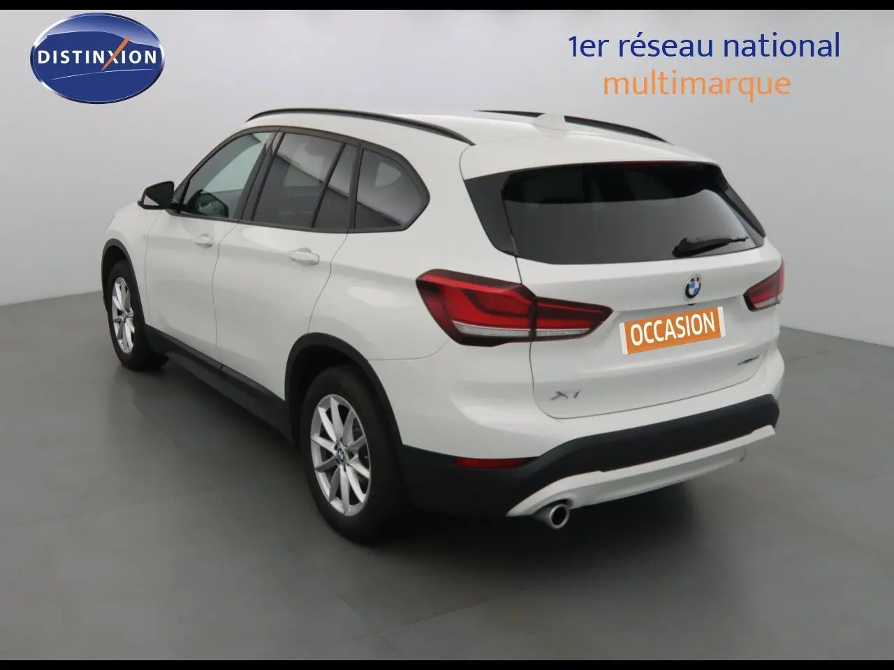 BMW X1 blanc Alpine métal vu en 3/4 arrière côté gauche dans un studio avec plaque occasion orange.