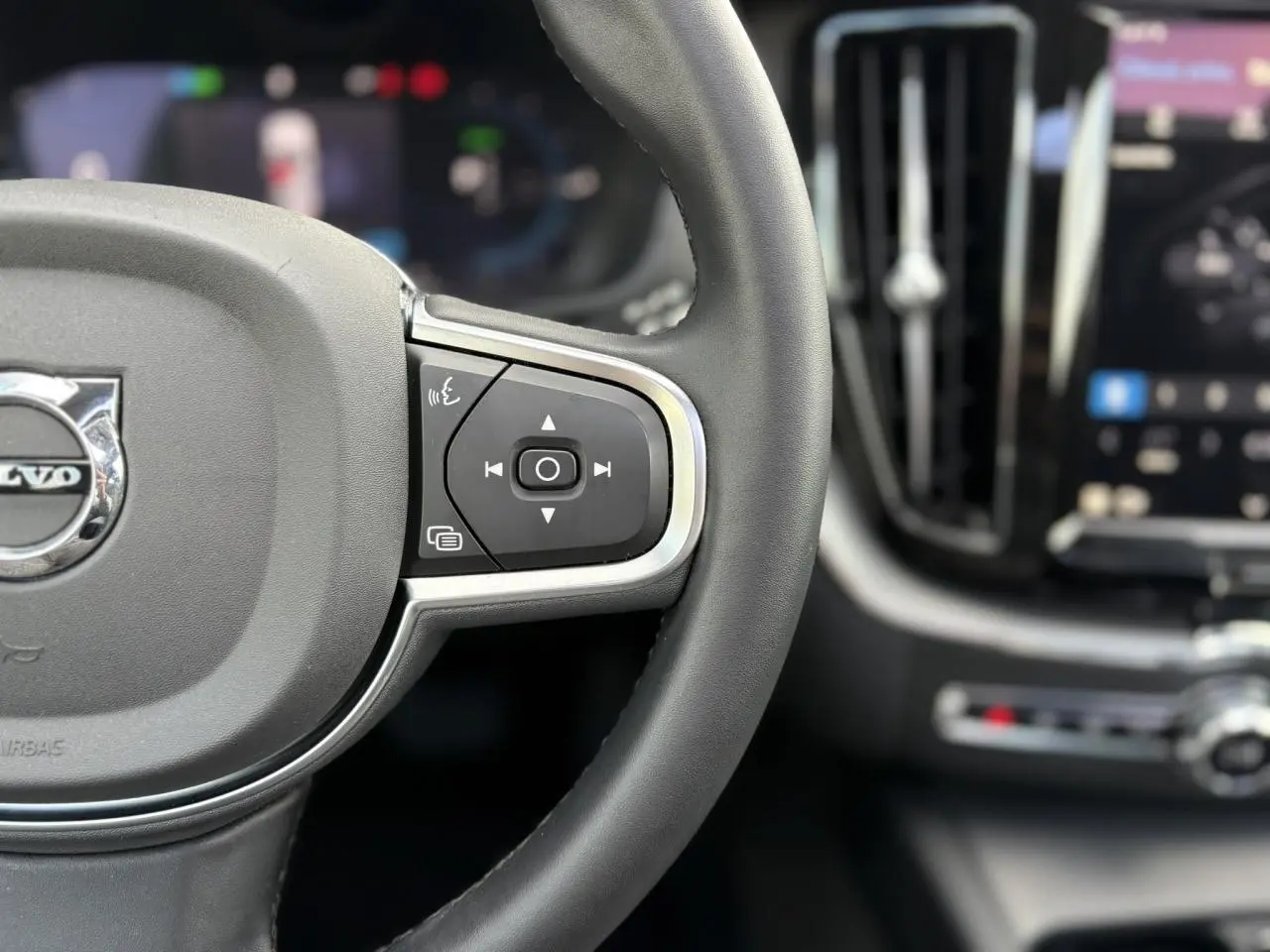Gros plan sur le volant noir en simili-cuir du Volvo XC60 2024 avec commandes intégrées et tableau de bord flou en arrière-plan.