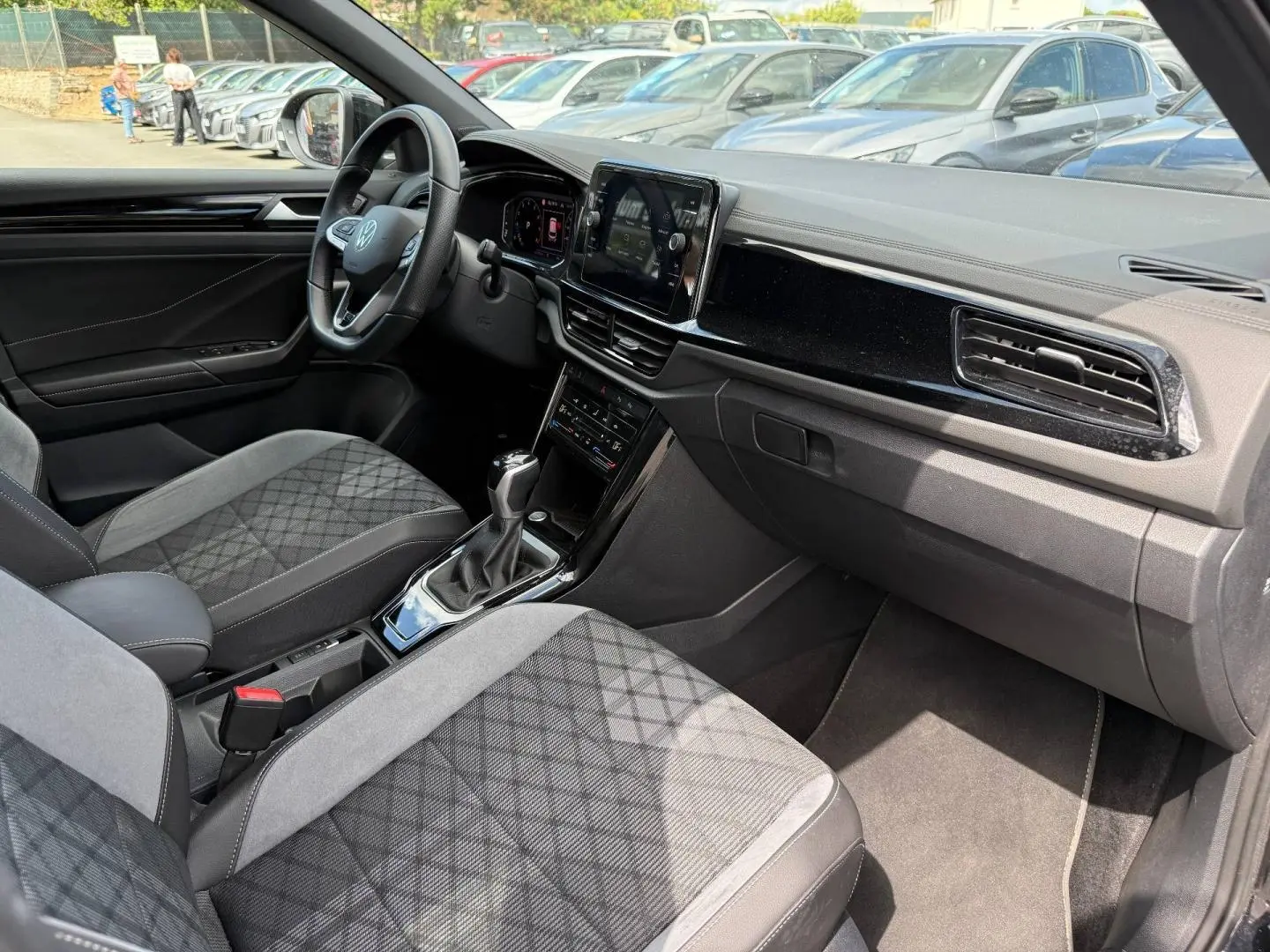 Vue intérieure côté conducteur du Volkswagen T-Roc 1.5 TSI R-Line 2025, sièges tissu gris et tableau de bord noir brillant.