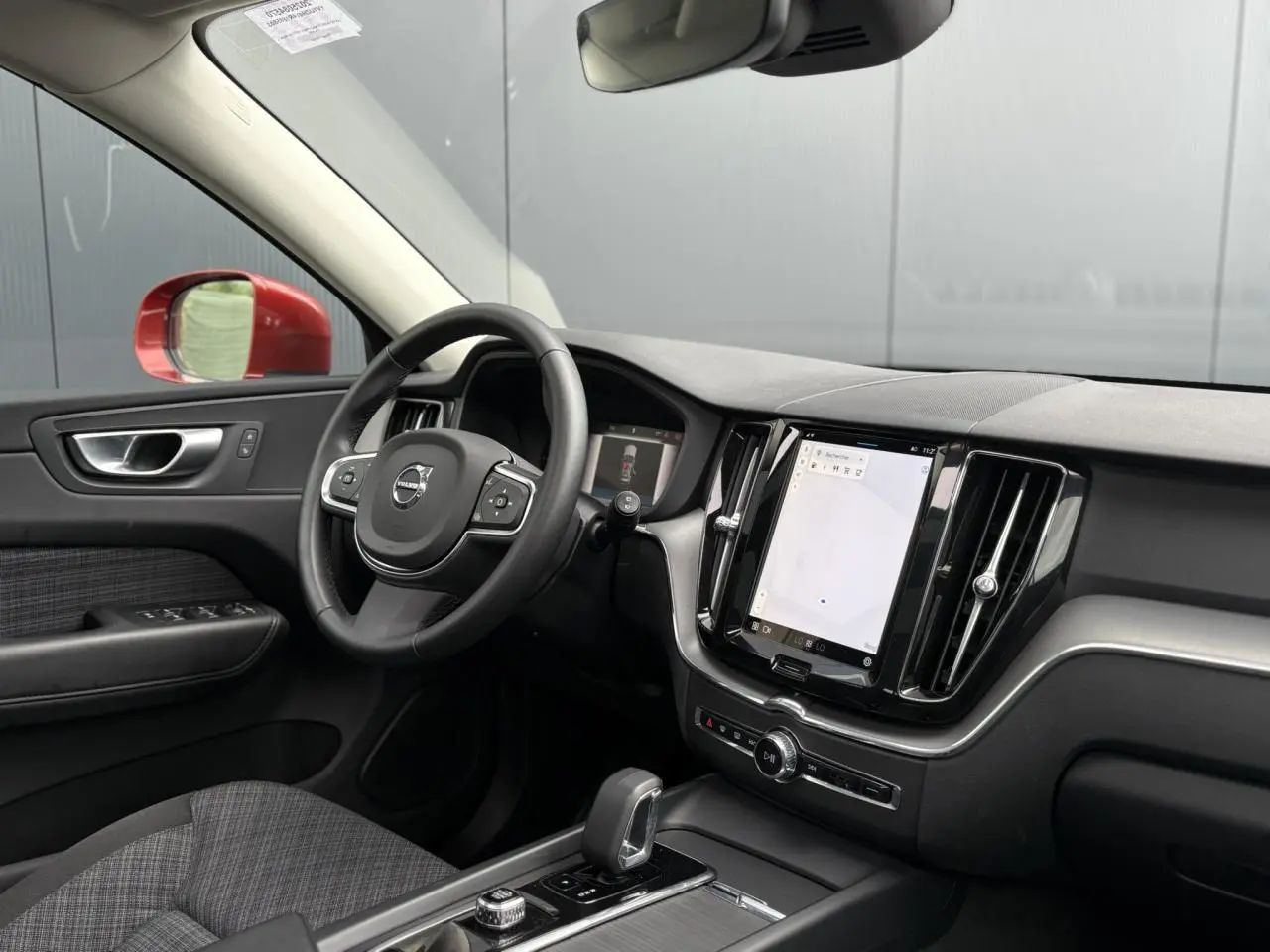 Vue intérieure côté conducteur du Volvo XC60 T6 AWD PHEV 2024, volant noir, écran tactile vertical et rétroviseur rouge visible.