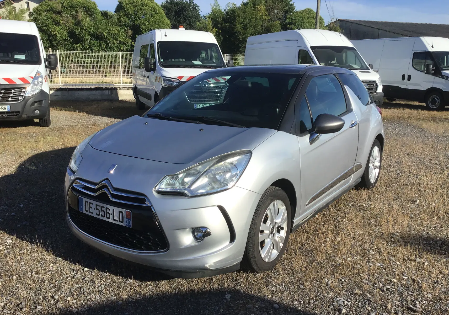 Citroën DS3 gris clair métal vue 3/4 avant droit sur un parking avec plusieurs fourgons blancs en arrière-plan.