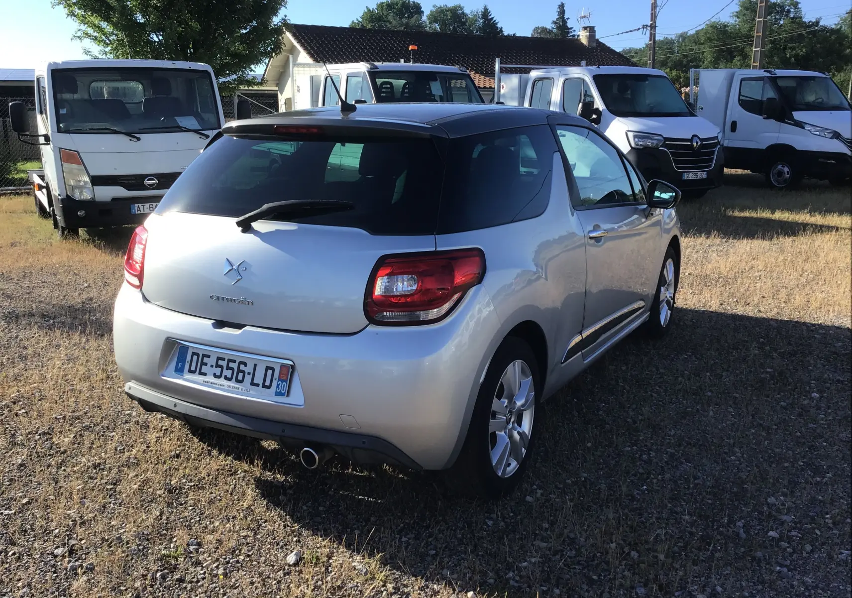 Vue 3/4 arrière droite d'une Citroën DS3 gris clair métal avec toit noir, garée sur un sol herbeux.