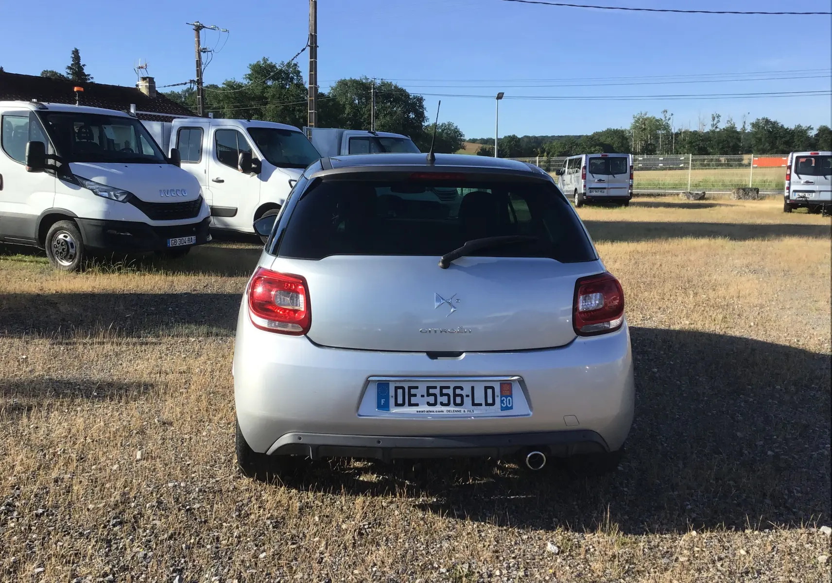 Vue arrière d'une Citroën DS3 gris clair métal, avec feux rouges et plaque d'immatriculation visible sur un terrain enherbé.