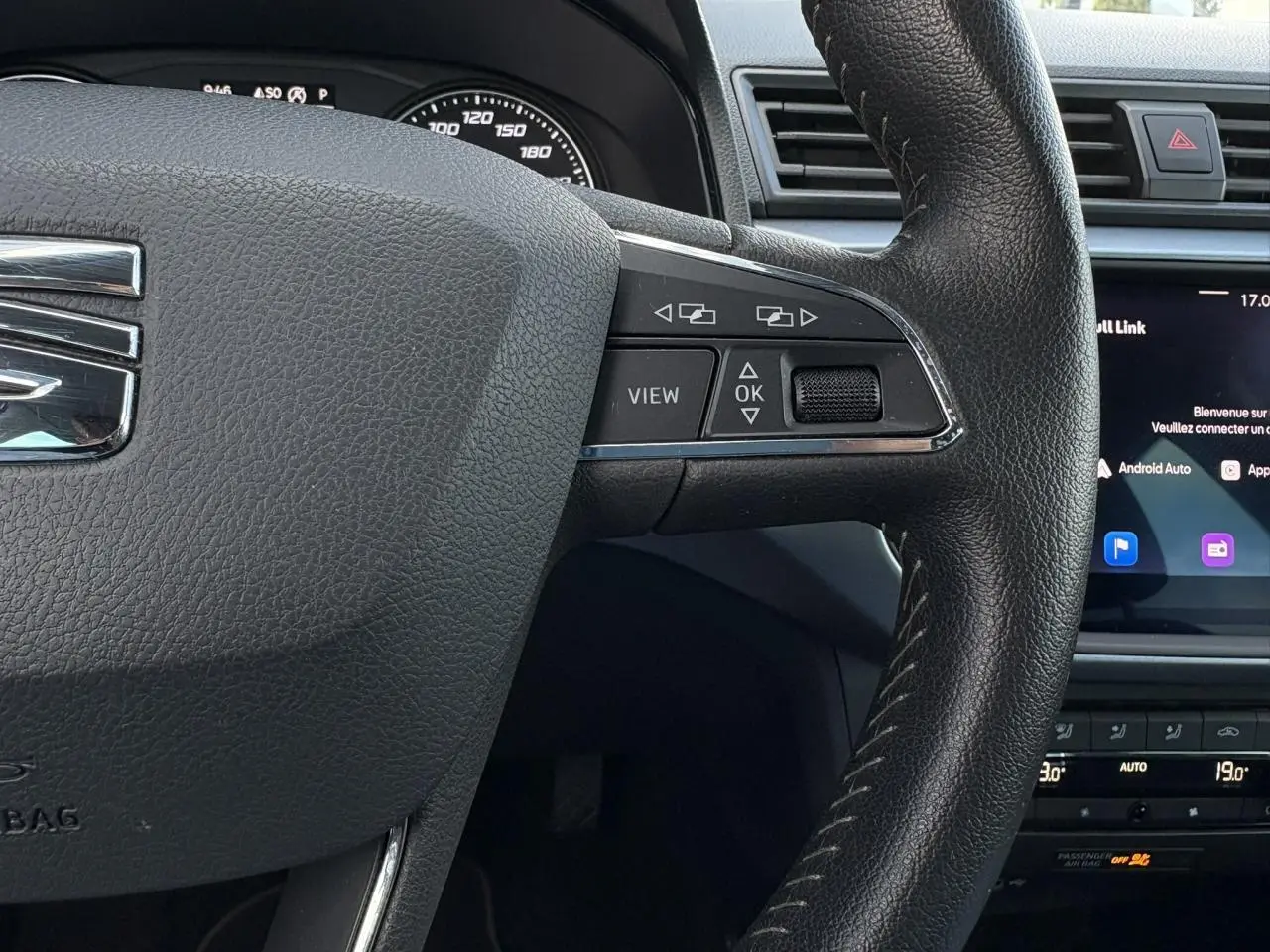 Gros plan sur le volant en cuir noir du SEAT Arona gris magnétique, avec commandes multifonctions et écran tactile en arrière-plan.