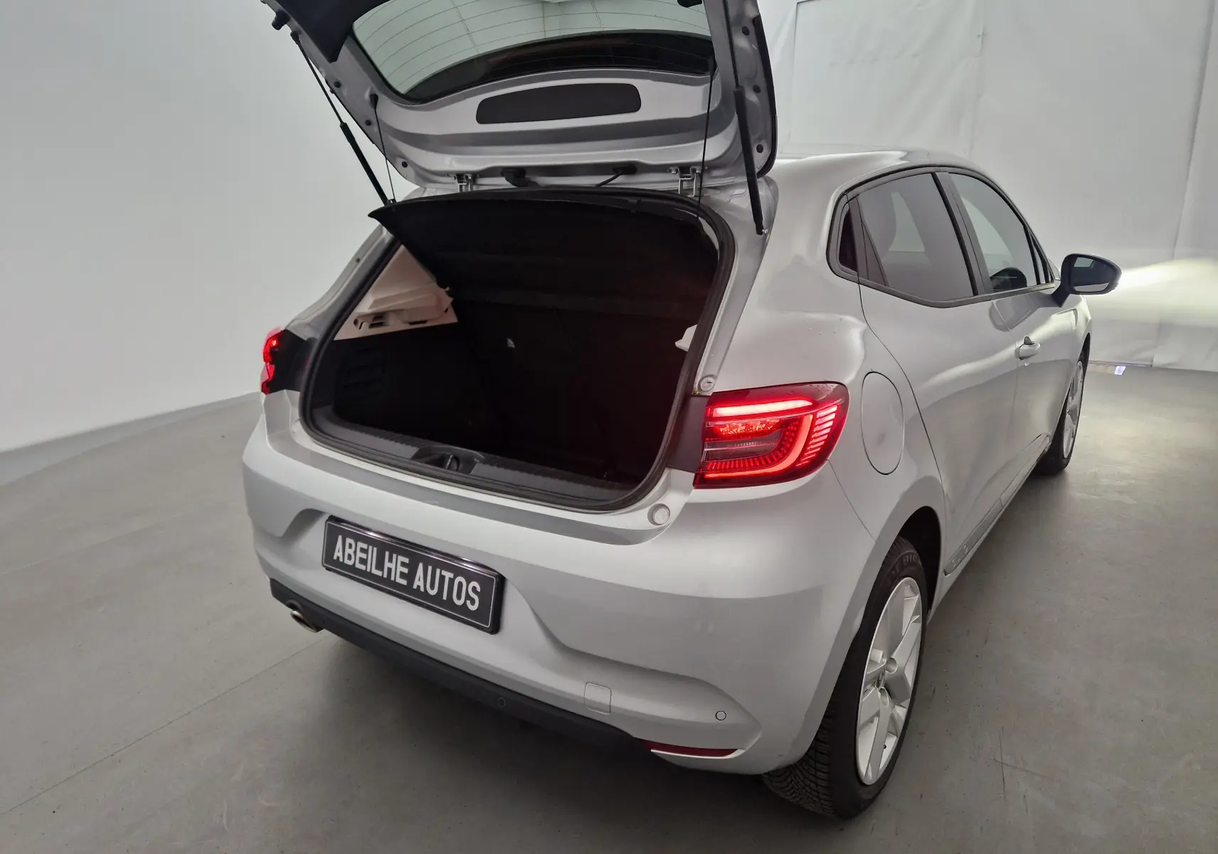 Renault Clio V gris platine vue 3/4 arrière droit avec coffre ouvert et feux arrière allumés dans un studio.