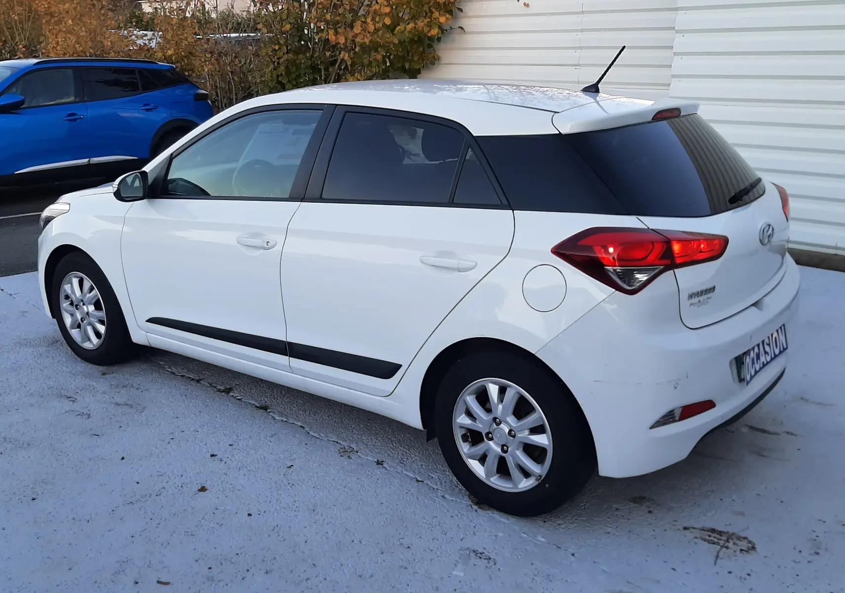 Hyundai i20 blanche vue en profil côté gauche, avec vitres arrière teintées et feux arrière allumés.