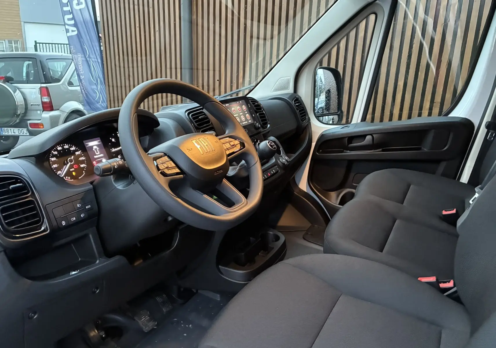 Vue intérieure côté conducteur du Fiat Ducato Fourgon blanc 2025, avec tableau de bord moderne et sièges tissu noir.