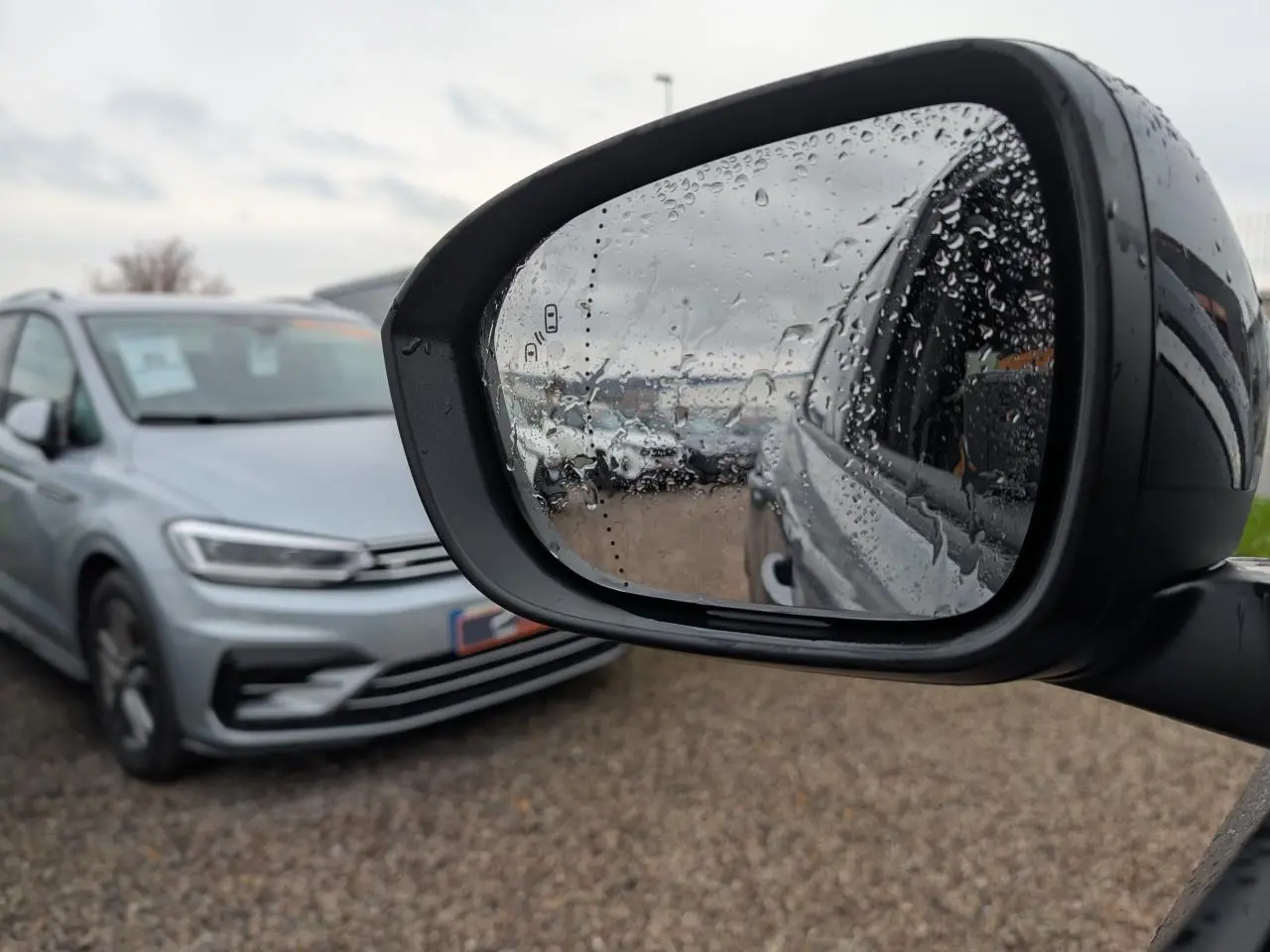 Rétroviseur côté droit avec gouttes d'eau reflétant un véhicule gris en stationnement, vue extérieure floue.