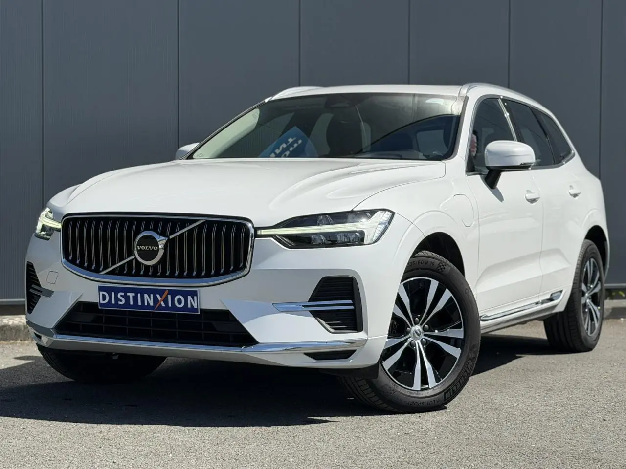 Volvo XC60 T6 AWD PHEV blanc Crystal en 3/4 avant droit, avec calandre chromée et jantes alliage 18 pouces.