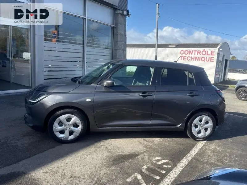 Profil droit d'une Suzuki Swift 1.2 Hybrid 83ch Privilège 2024 en Mineral Gray Metal, garée devant un bâtiment.