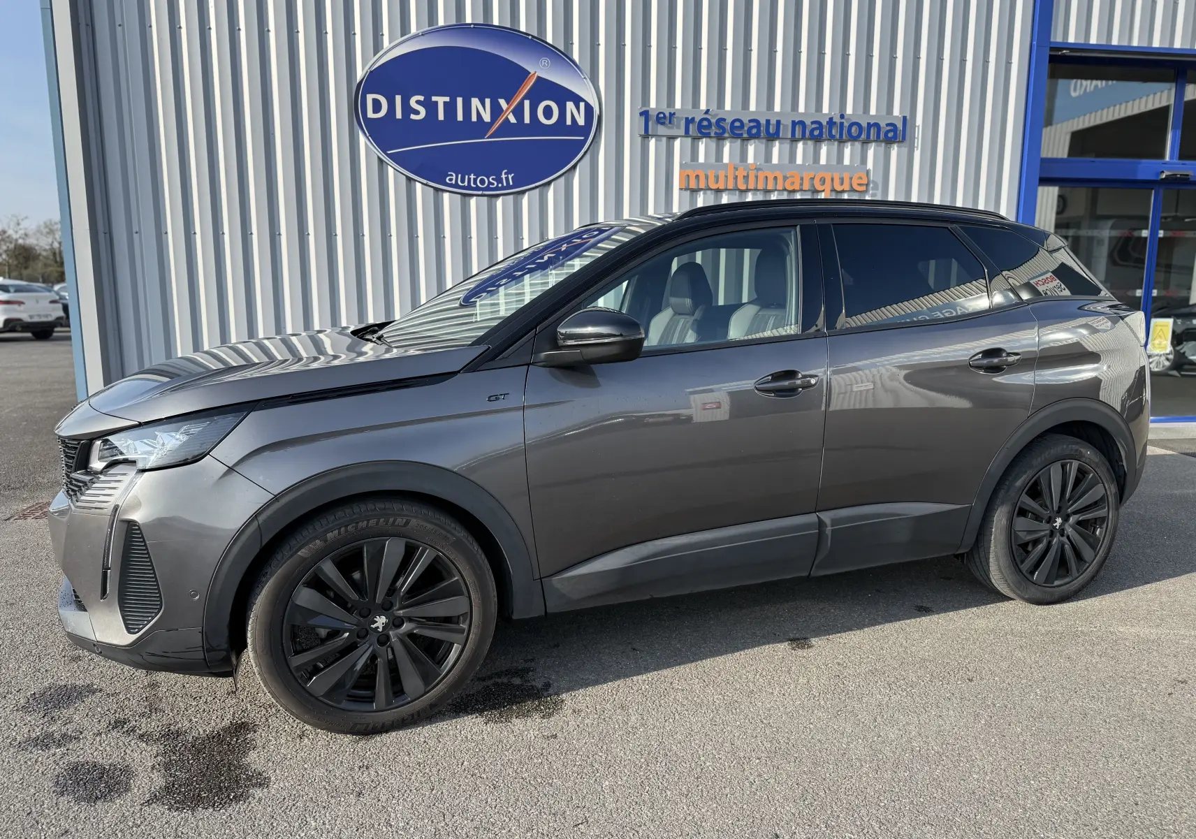 Peugeot 3008 gris en profil droit, finition GT avec jantes noires et toit noir, stationné devant un bâtiment commercial.