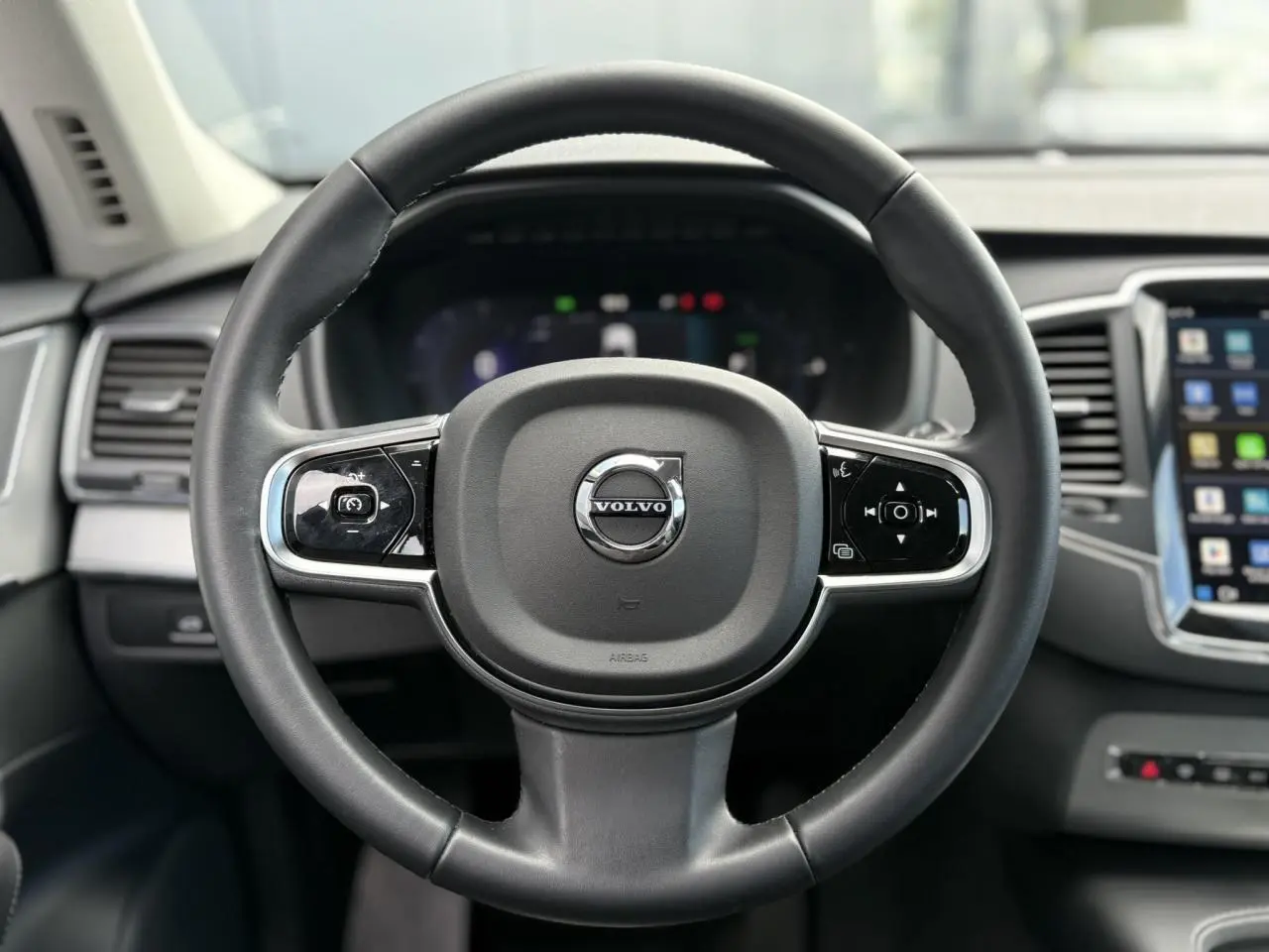 Vue rapprochée du volant noir du Volvo XC90 T8 2024 avec commandes intégrées et tableau de bord digital flou en arrière-plan.