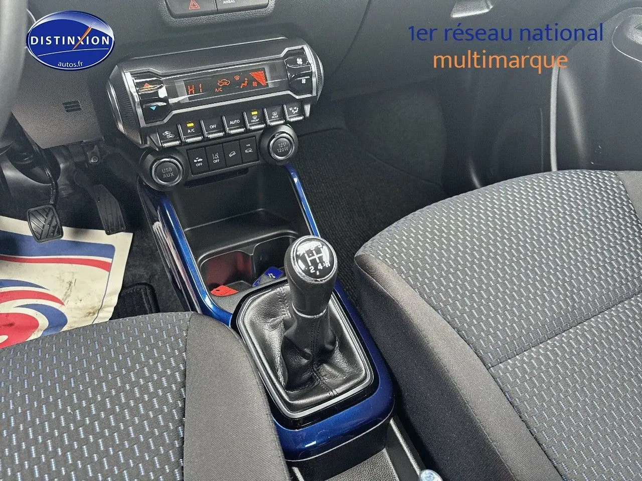Vue intérieure centrée sur la console centrale et levier de vitesse manuelle, sièges tissu gris avec motifs bleus.
