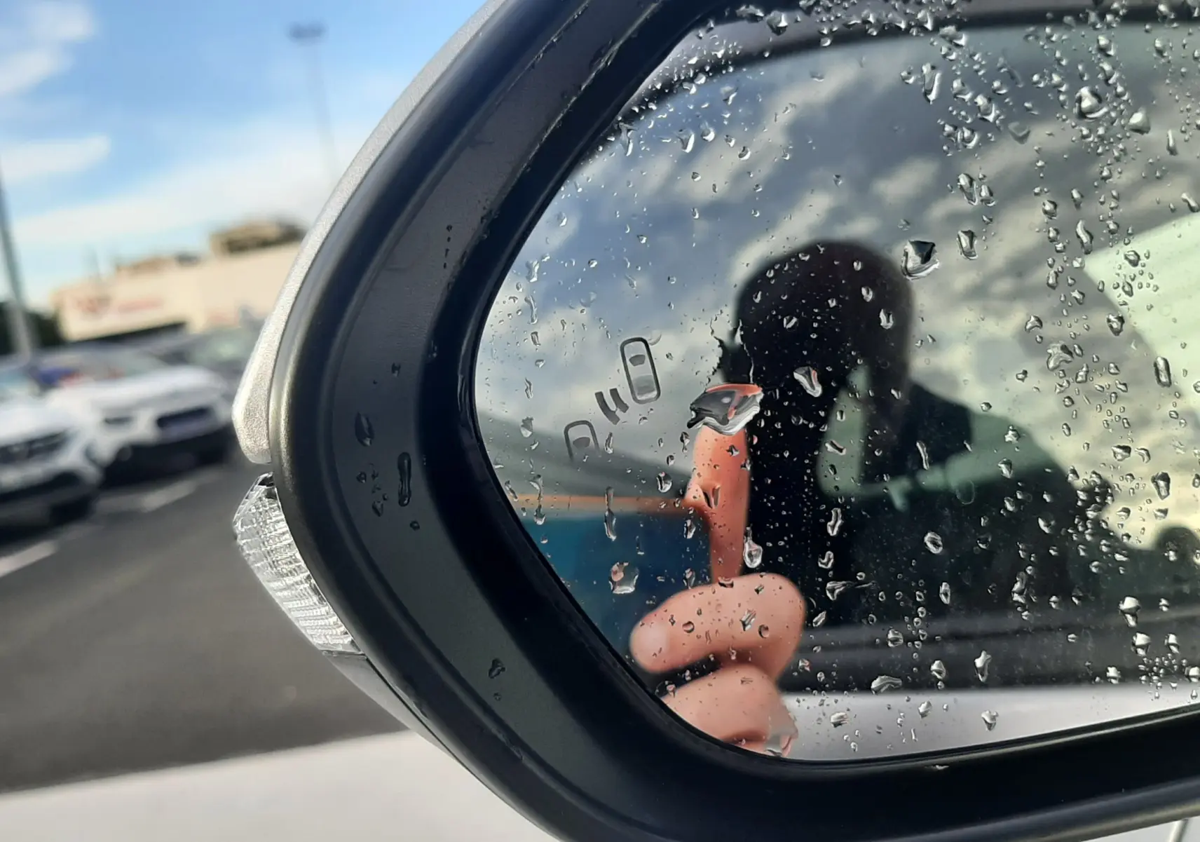 Rétroviseur extérieur droit d'une Toyota Yaris Cross gris minéral, avec gouttes d'eau et reflet flou d'une personne.