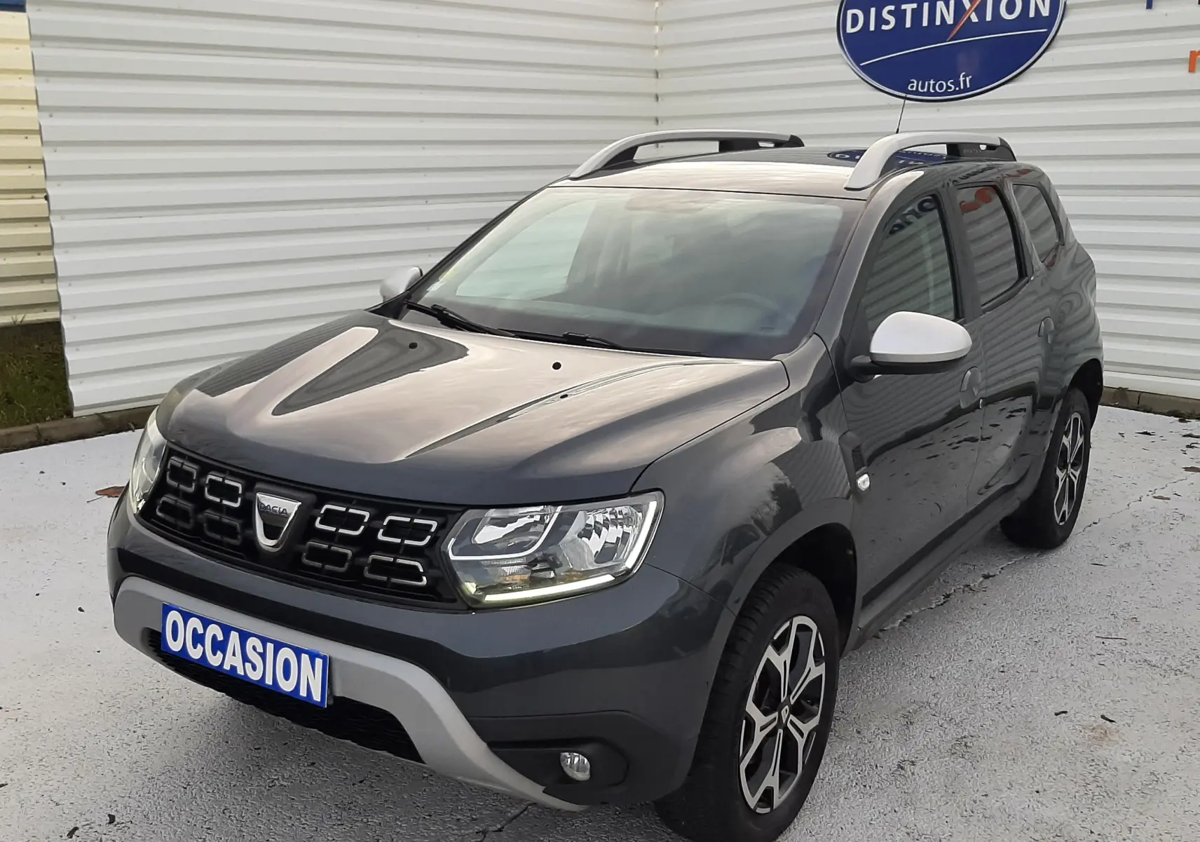 Dacia Duster gris Cassiopée en 3/4 avant droit, avec jantes bi-ton et barres de toit visibles.