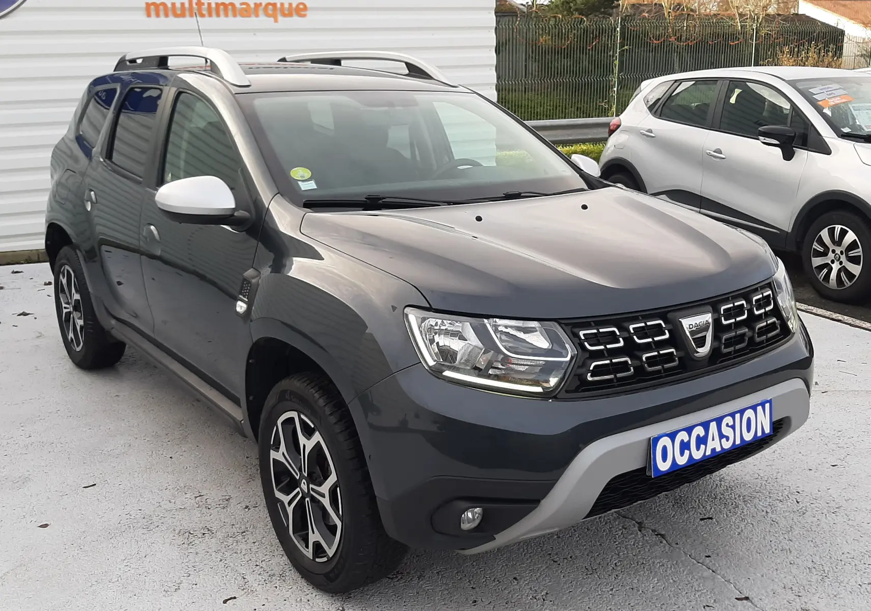 Dacia Duster gris cassiopée en 3/4 avant droit, avec jantes bi-ton et barres de toit visibles sur parking.