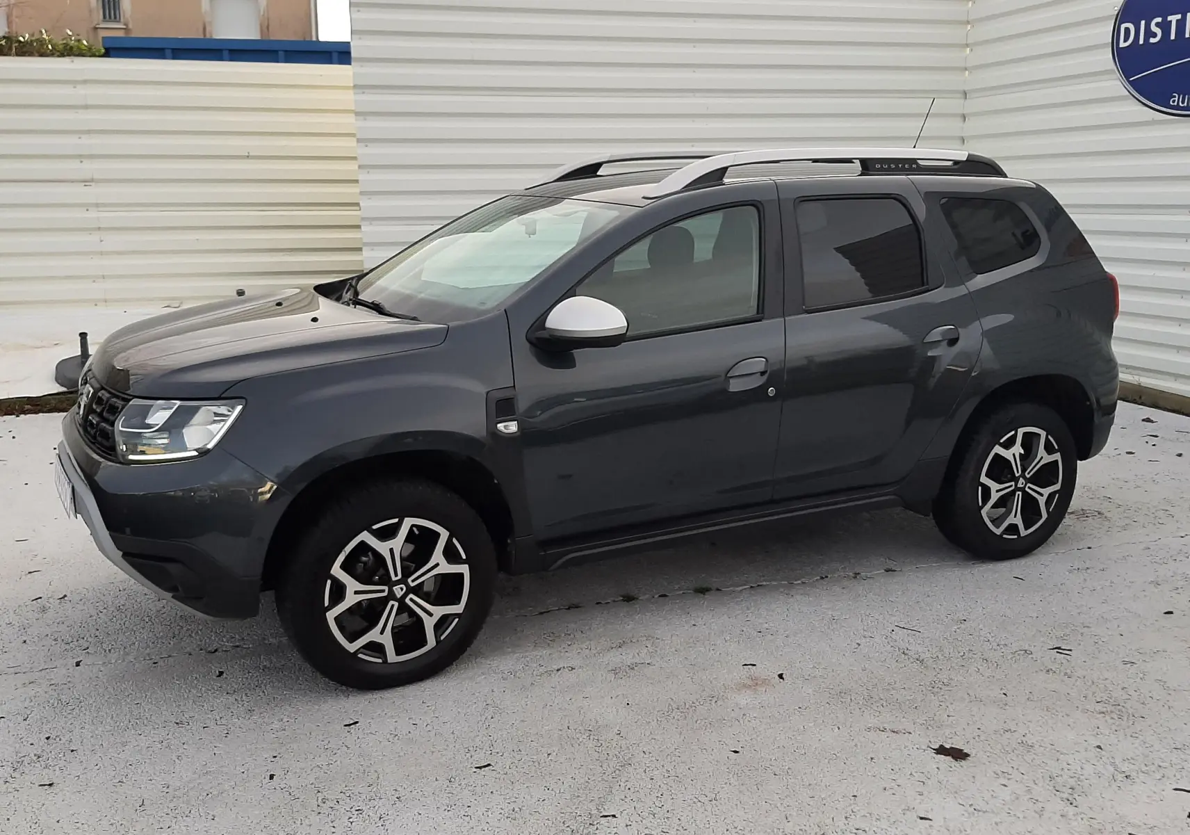 Vue latérale gauche d'un Dacia Duster gris Cassiopée 2019 avec jantes alliage bicolores et barres de toit.