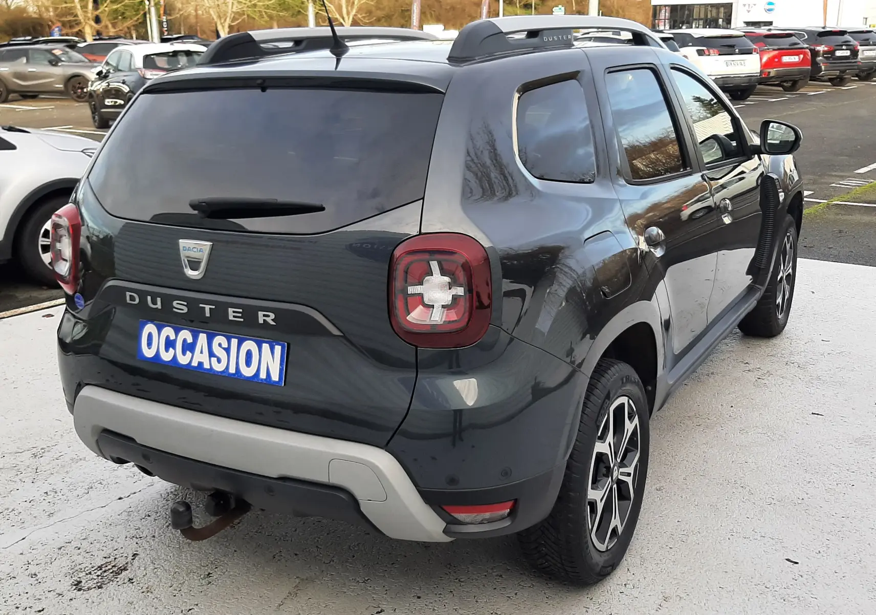 Vue 3/4 arrière droite d'un Dacia Duster gris Cassiopée avec barres de toit et attelage visible sur parking.
