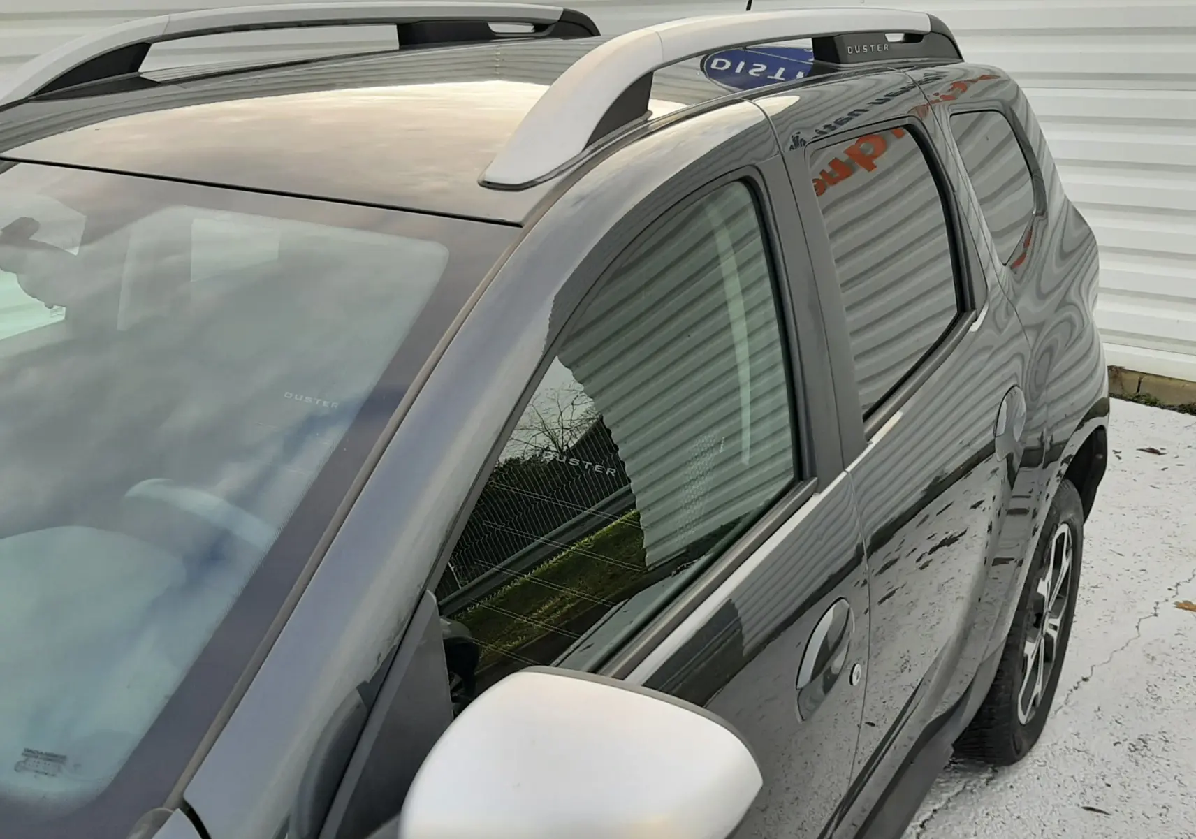 Vue 3/4 avant droit du Dacia Duster gris Cassiopée, avec barres de toit blanches et rétroviseur blanc contrastant.