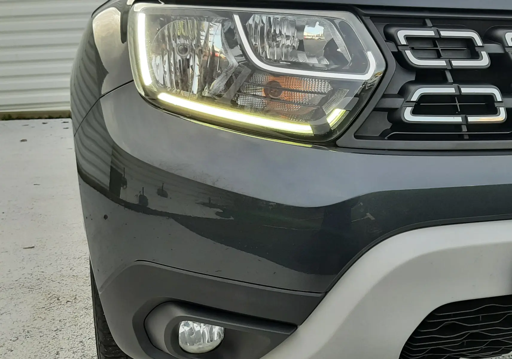 Gros plan sur l'avant droit du Dacia Duster gris Cassiopée, mettant en valeur le phare LED et la calandre noire.
