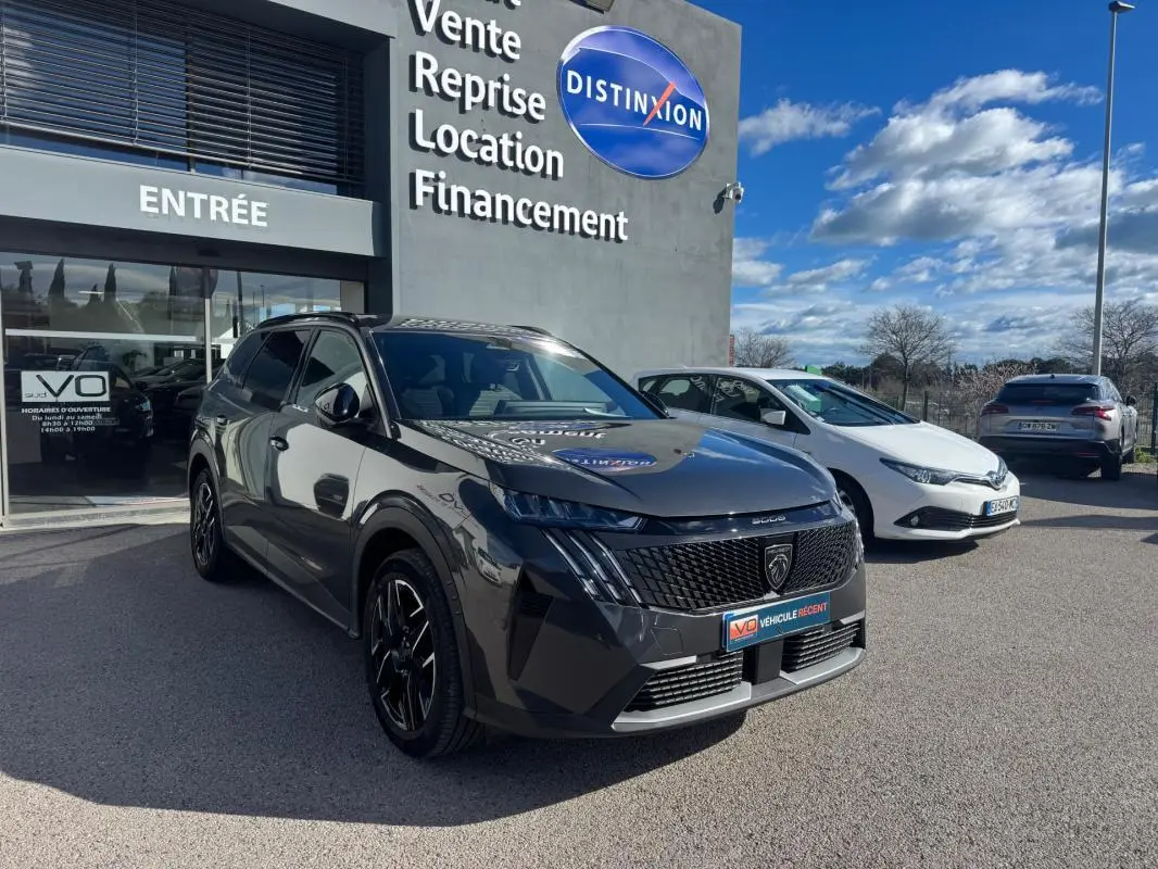Peugeot 5008 Hybrid gris en 3/4 avant droit, avec jantes alliage noires et calandre distinctive, devant un showroom.