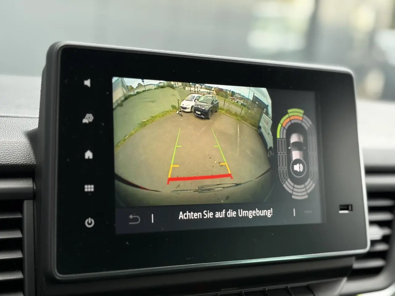 Écran tactile affichant la caméra de recul avec vue arrière sur un parking, dans un Nissan Primastar noir métal.