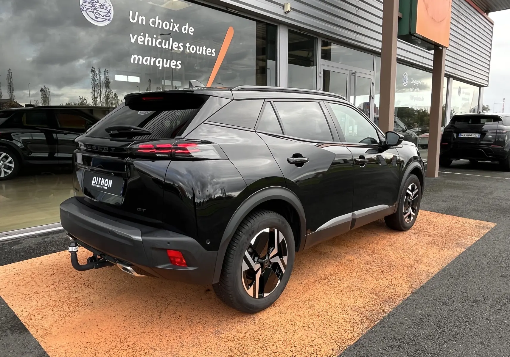 Peugeot 2008 noir en 3/4 arrière droit, avec jantes distinctives et attelage visible, stationné devant un showroom.