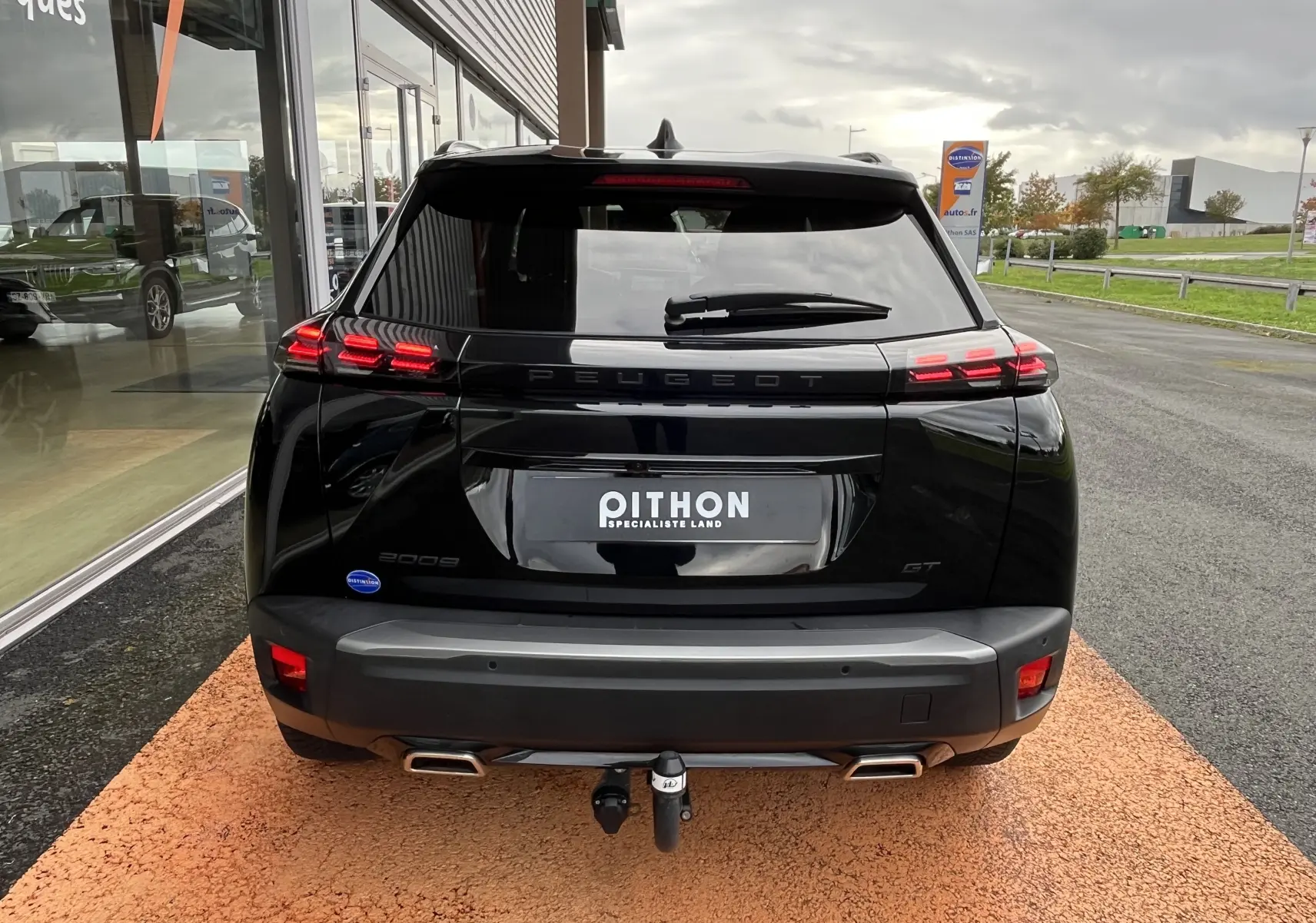 Vue arrière d’un Peugeot 2008 noir 2024 GT avec feux LED allumés et attelage visible sur un parking extérieur.