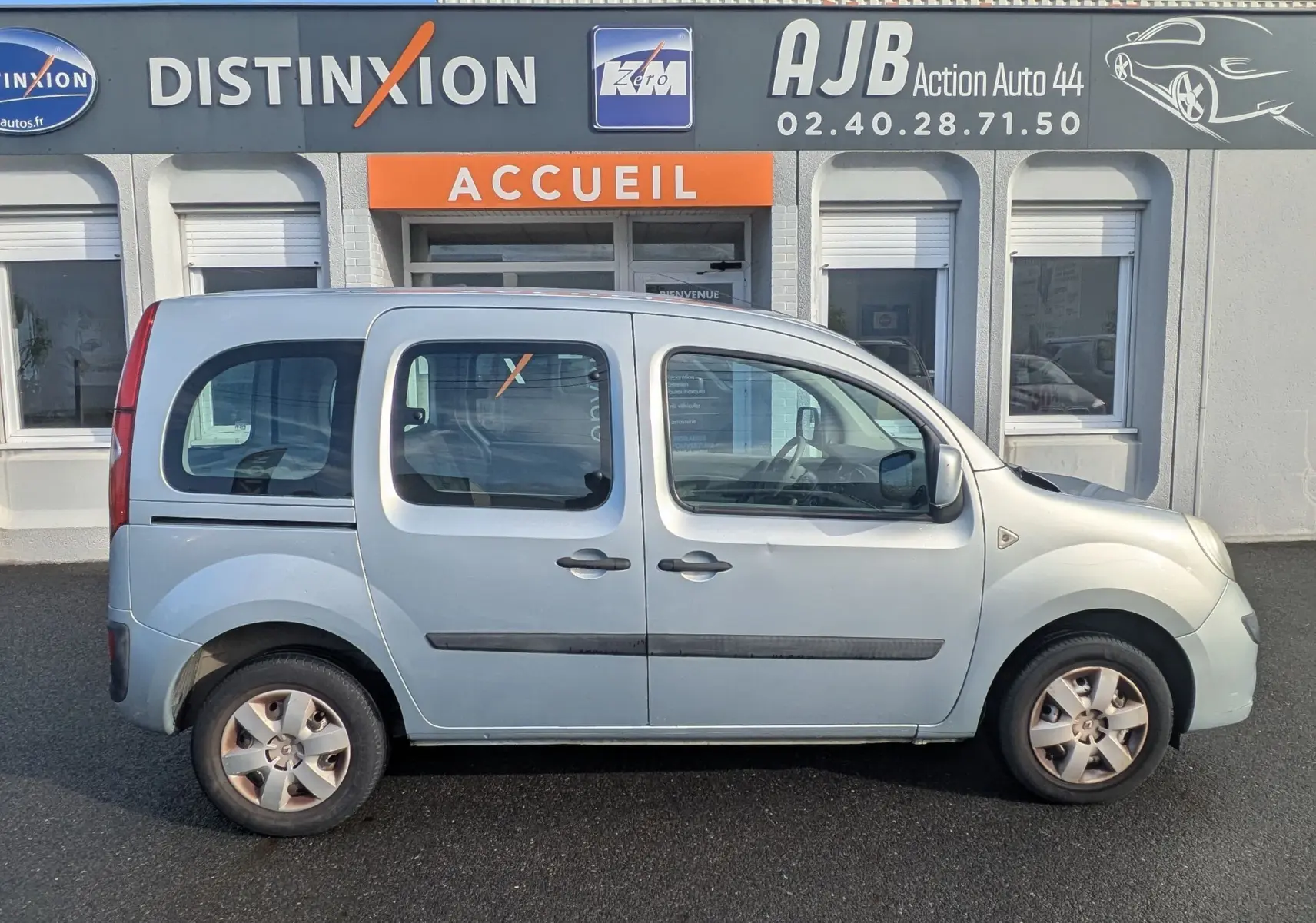 Profil latéral droit d'un Renault Kangoo II bleu ciel stationné devant un bâtiment commercial avec portes coulissantes visibles.