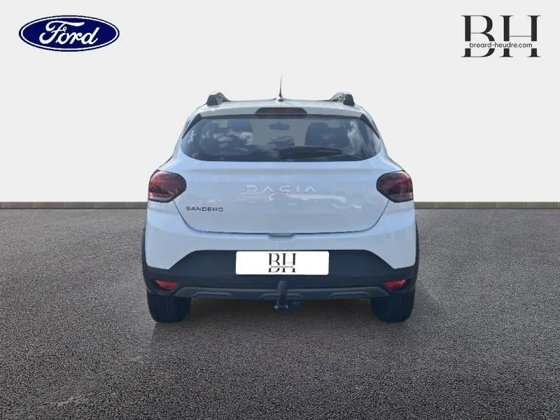 Vue arrière d'une Dacia Sandero blanche avec attelage et barres de toit visibles sur un fond neutre.