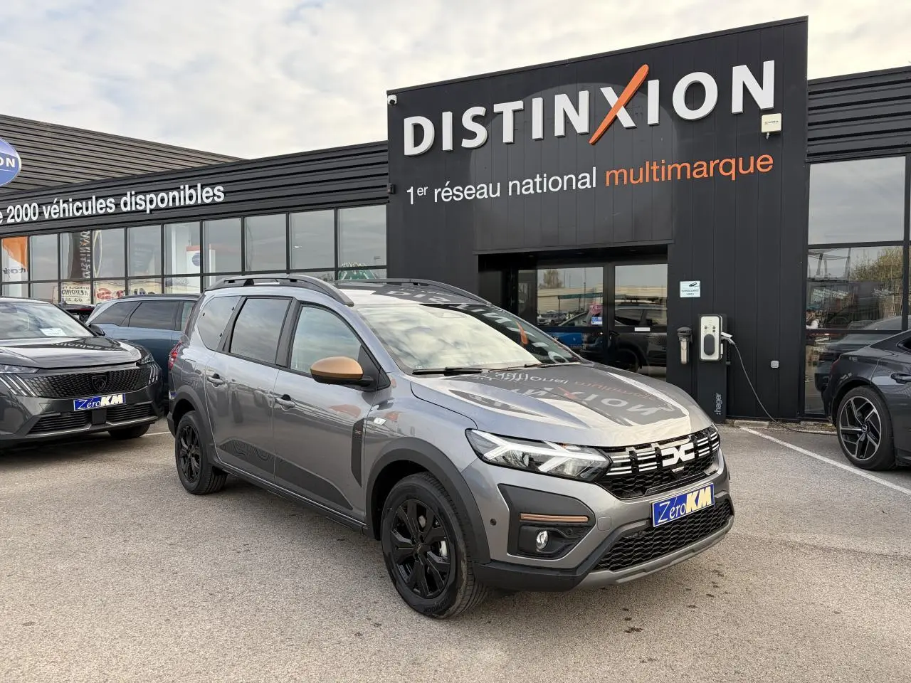 Vue 3/4 avant du Dacia Jogger gris schiste métal avec calandre noire et jantes aluminium noires devant un showroom Distinxion.