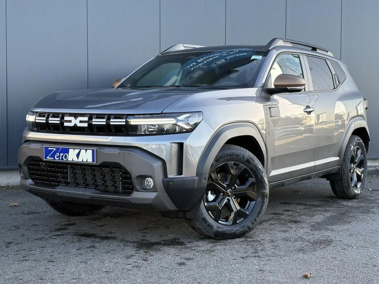 Vue 3/4 avant droite du Dacia Duster 2026 gris schiste avec jantes alliage noires et barres de toit longitudinales.