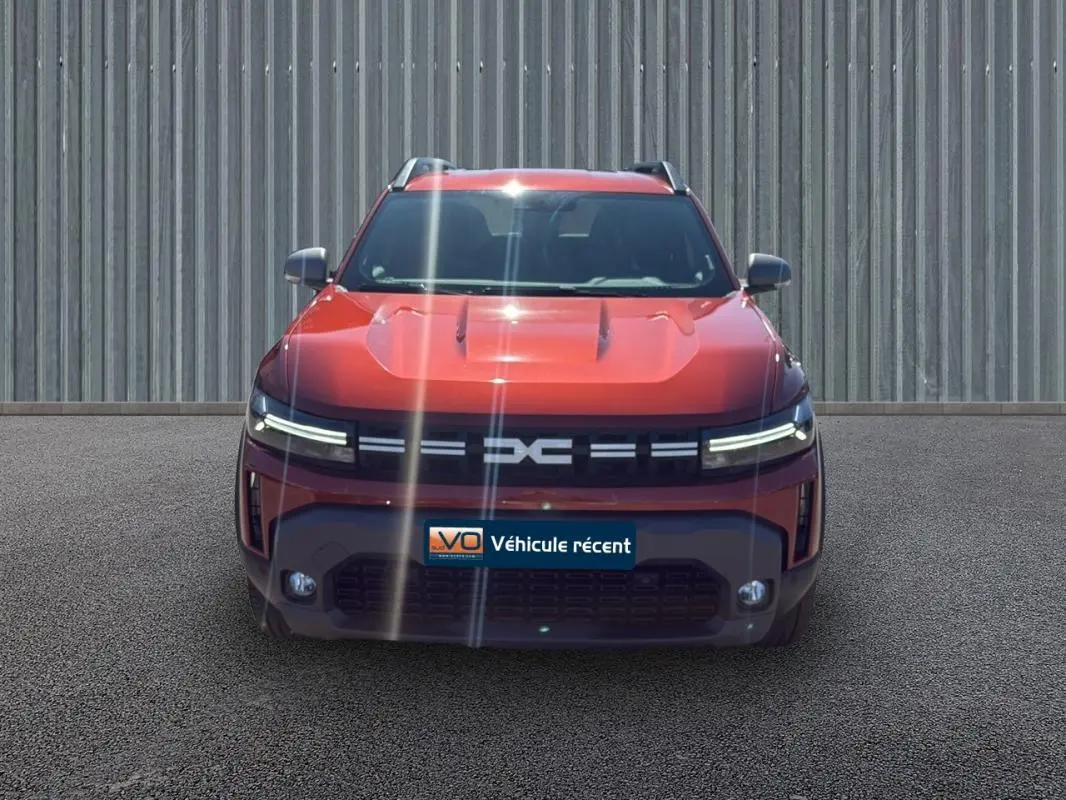 Vue frontale du Dacia Duster 2025 orange avec phares LED fins et barres de toit visibles sur fond urbain.