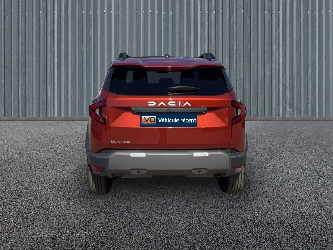 Vue arrière du Dacia Duster orange 2025 avec vitres surteintées et barres de toit visibles sur fond industriel.