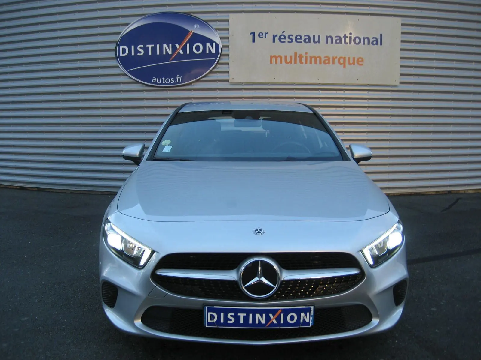 Vue frontale d'une Mercedes Classe A180D gris métallisé avec phares LED allumés, devant un mur avec logo Distinxion.