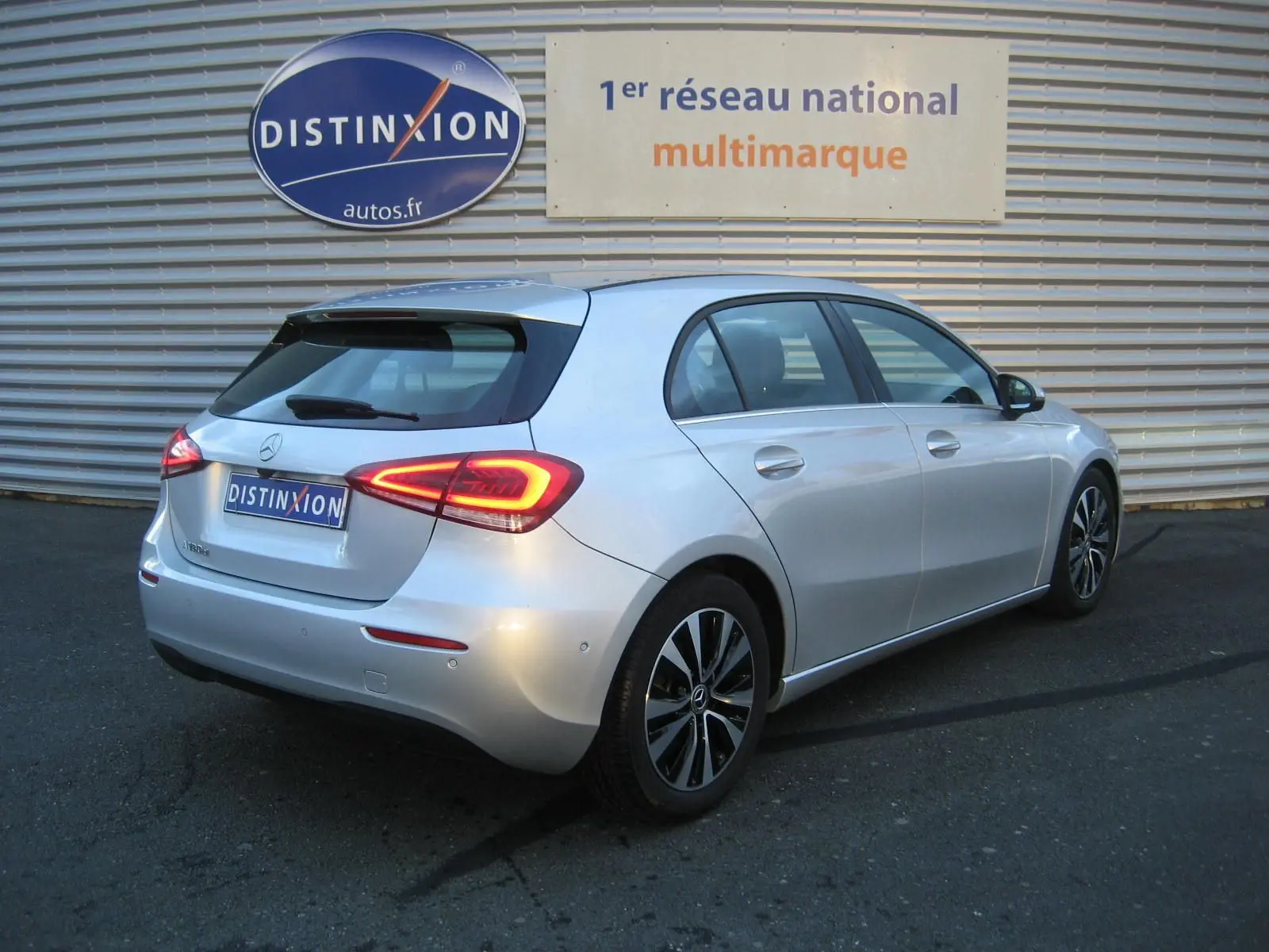 Vue 3/4 arrière droite d'une Mercedes Classe A180D gris métallisé avec feux LED allumés, garée devant un mur métallique.