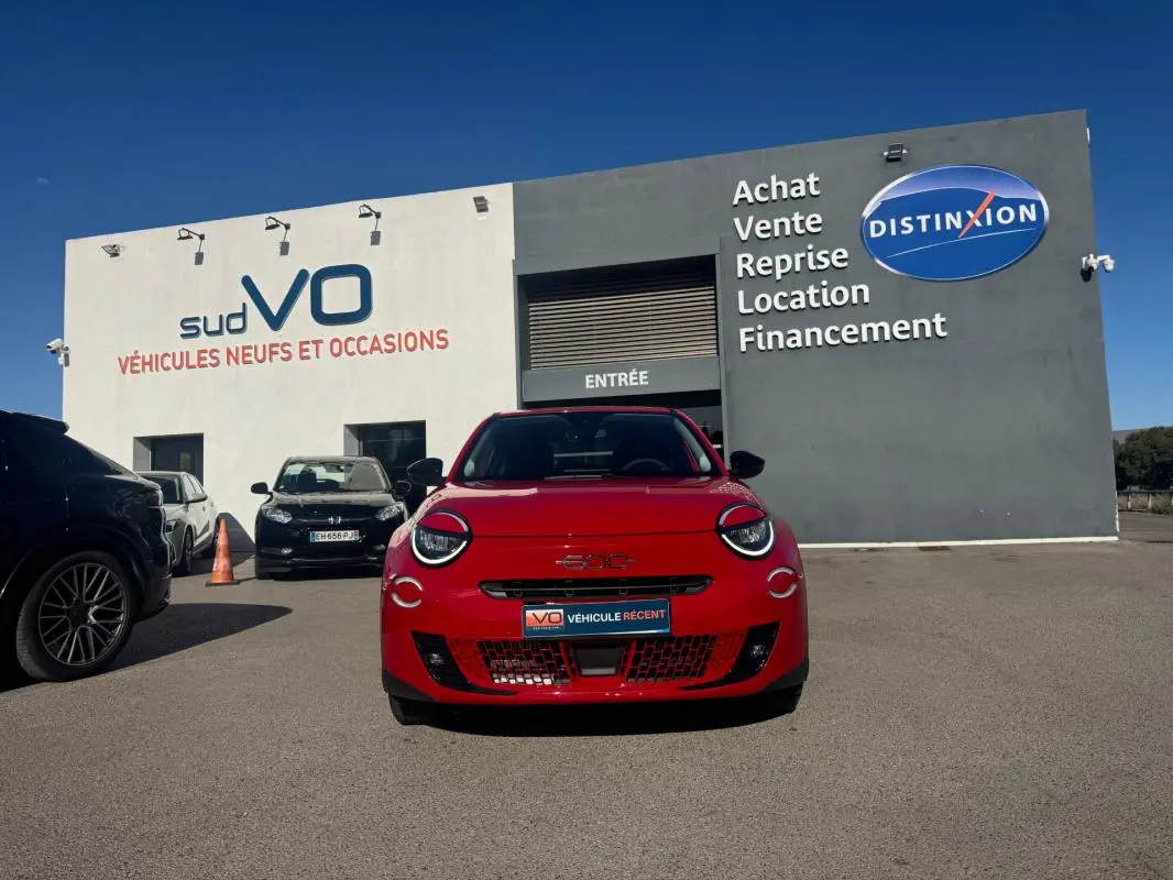 Vue frontale d'une FIAT 600 T-Gen 3 rouge 2025, avec phares ronds et calandre ajourée, devant un garage de véhicules d'occasion.