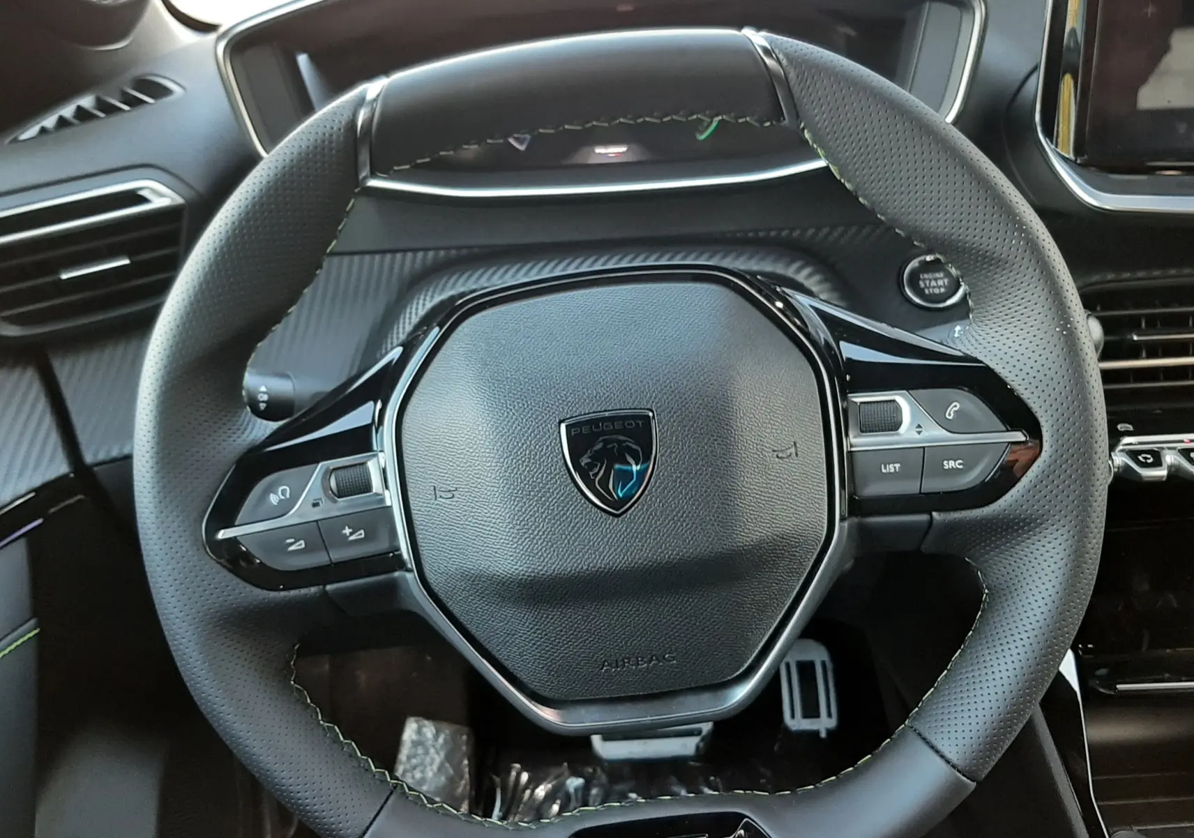 Vue rapprochée du volant cuir noir avec logo Peugeot et badge GT, tableau de bord et commandes intégrées visibles.