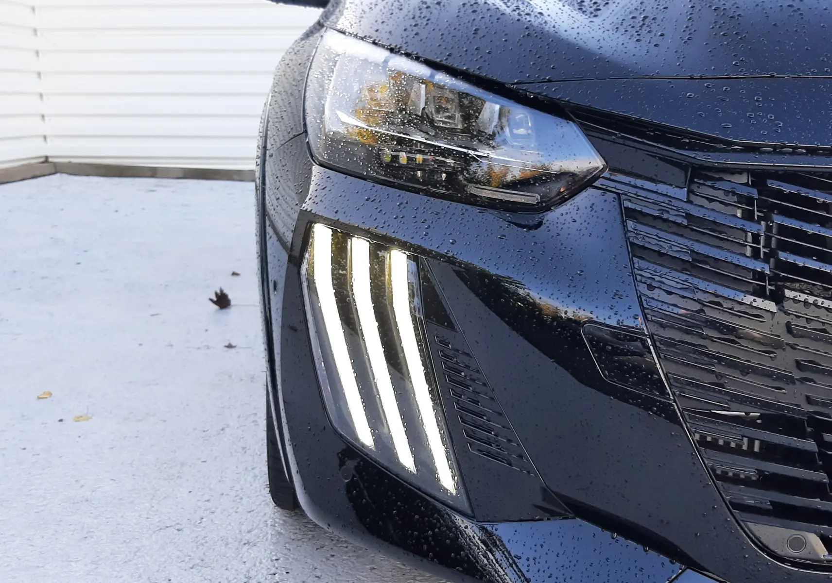 Gros plan sur l'avant gauche noir perla d'une Peugeot 208 Hybrid 145ch GT avec feux LED verticaux allumés sous la pluie.