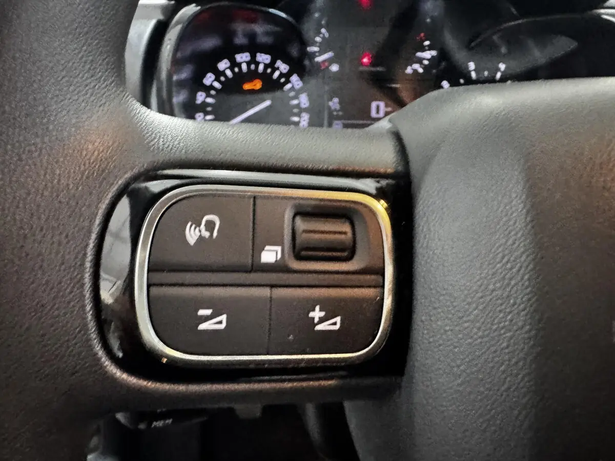 Gros plan sur les commandes gauche du volant multifonctions noir d'une Citroën C3 blanche, avec tableau de bord en arrière-plan flou.