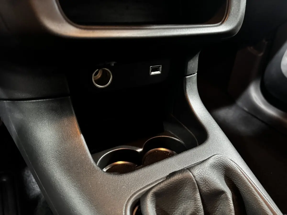 Gros plan sur la console centrale noire de la Citroën C3 avec prise 12V, port USB et emplacement porte-gobelets.