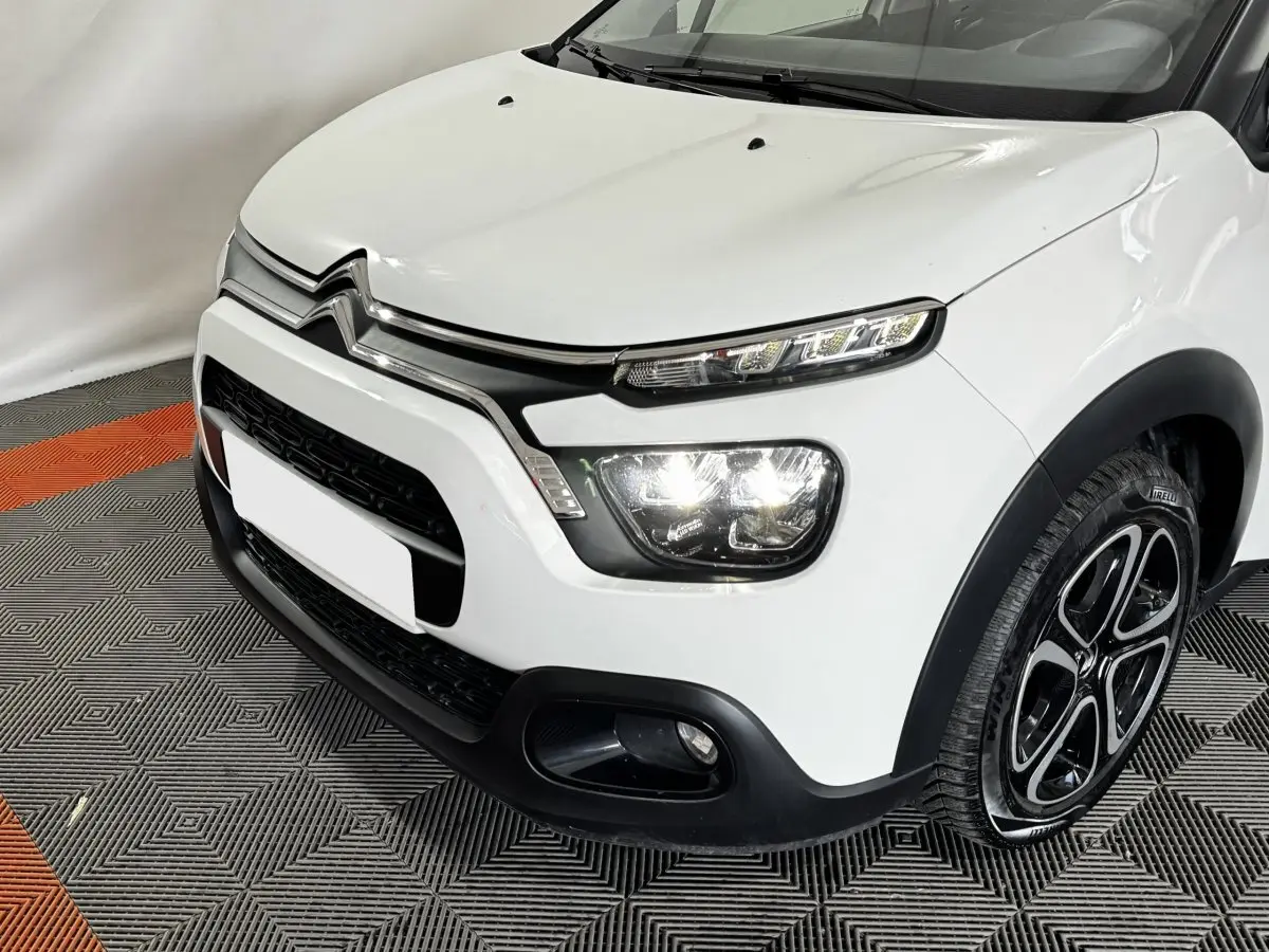 Vue 3/4 avant droit d'une Citroën C3 blanche 2024 avec phares LED allumés et jantes noires à motifs.