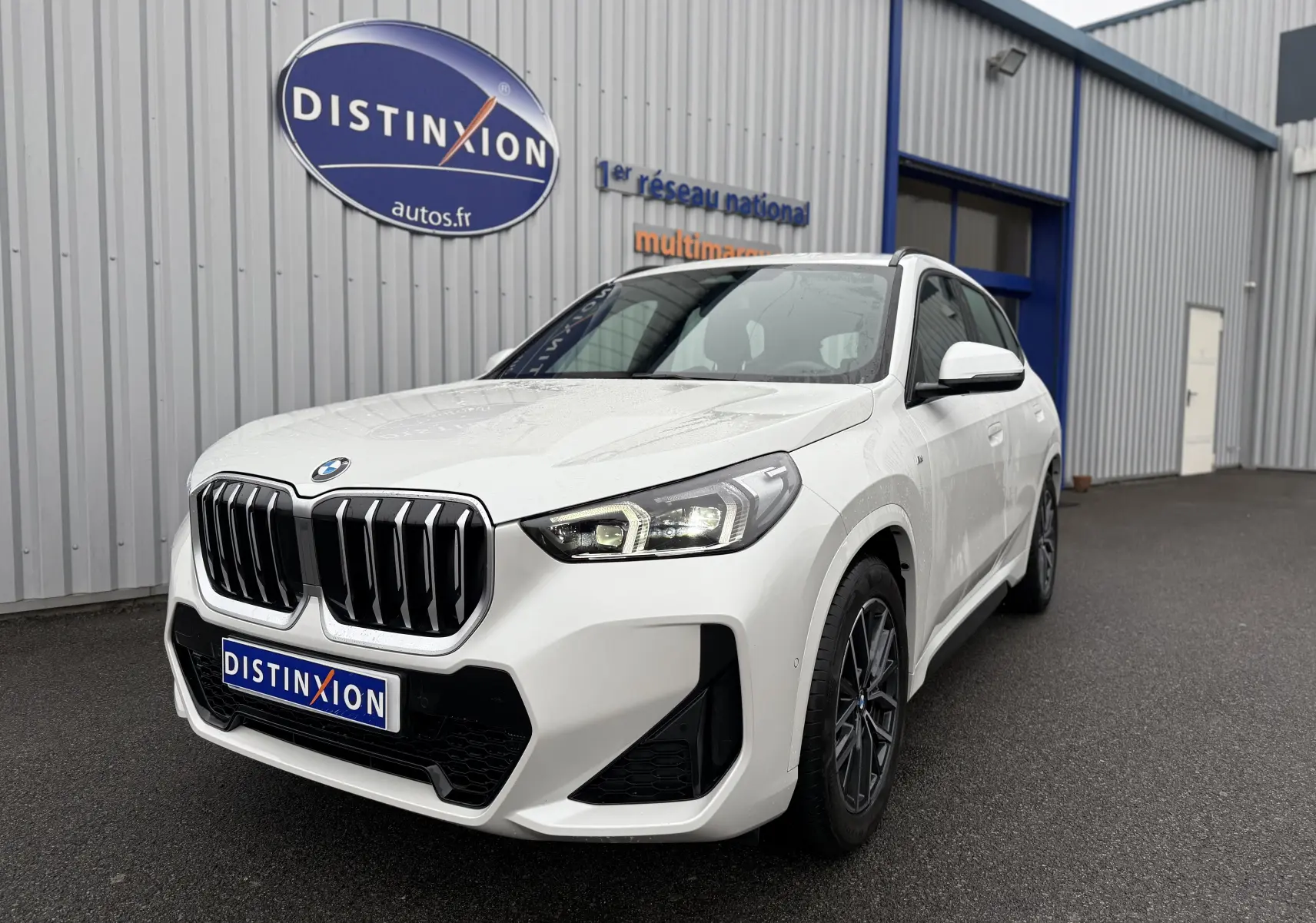 BMW X1 blanc minéral en 3/4 avant droit, avec calandre noire et phares LED distinctifs, stationné devant un bâtiment.