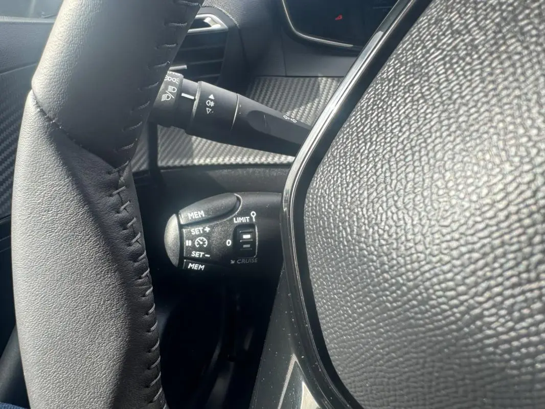 Gros plan sur la commande de régulateur de vitesse à gauche du volant dans l’habitacle noir du Peugeot 2008 blanc 2024.