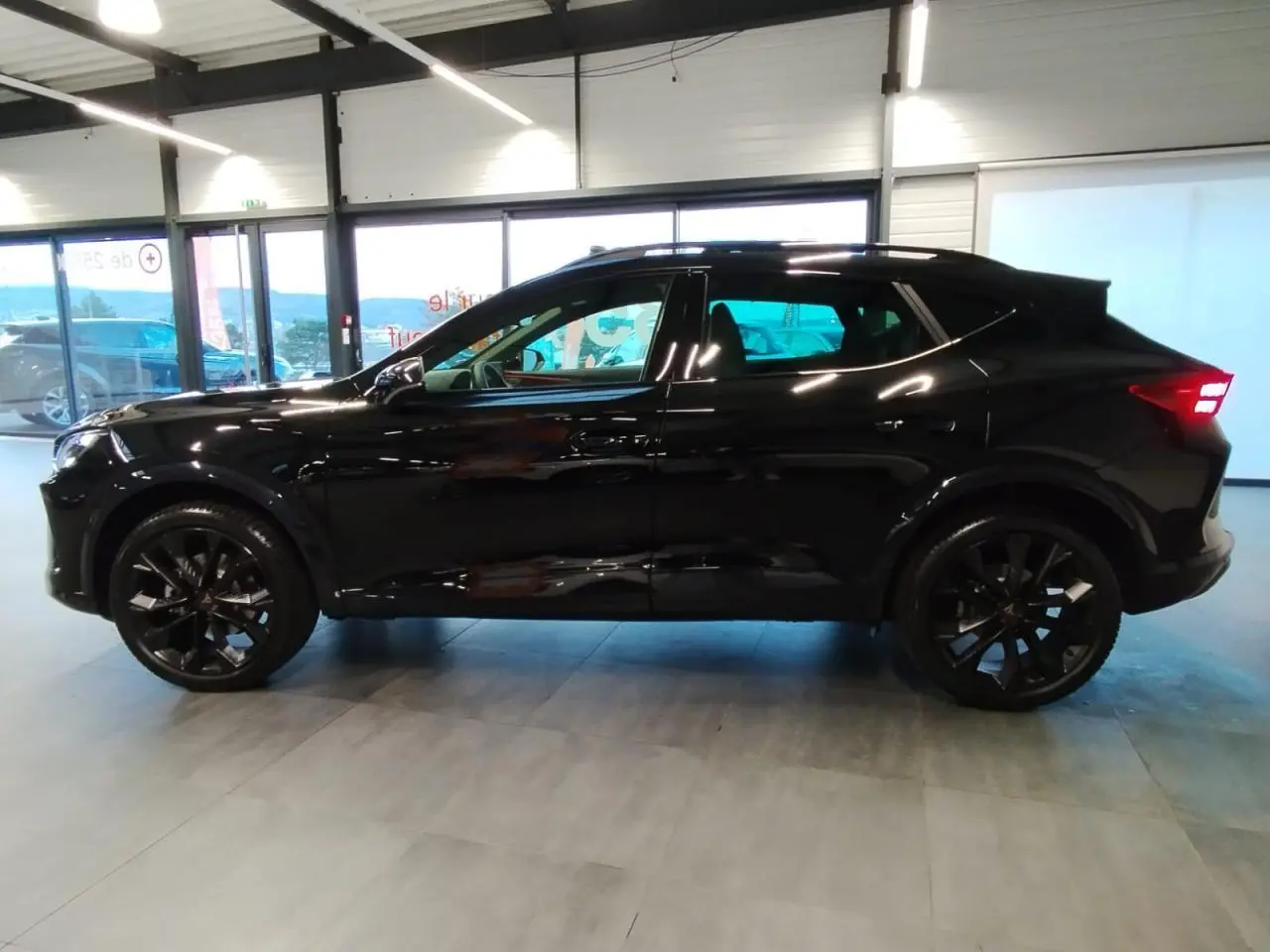 Vue de profil côté gauche d’un CUPRA Formentor noir métal avec jantes alliage noires et feux arrière allumés en intérieur showroom.